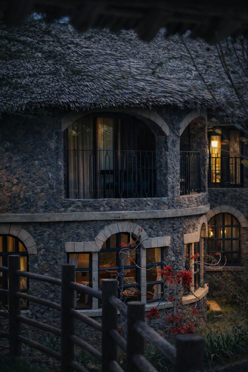 O'Leleshwa villa at dusk — stone facade and arched balcony, one window lamplit, coral-tree blooms in the foreground.
