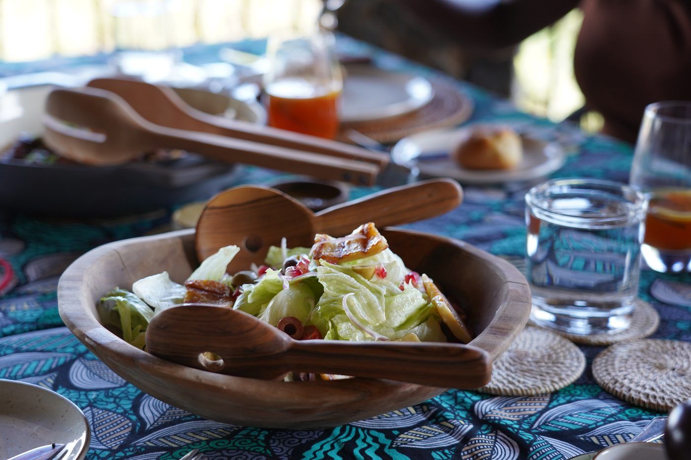 A salad of grilled halloumi, olives and pomegranate in an olive-wood bowl with carved servers, on a layered blue kanga table.