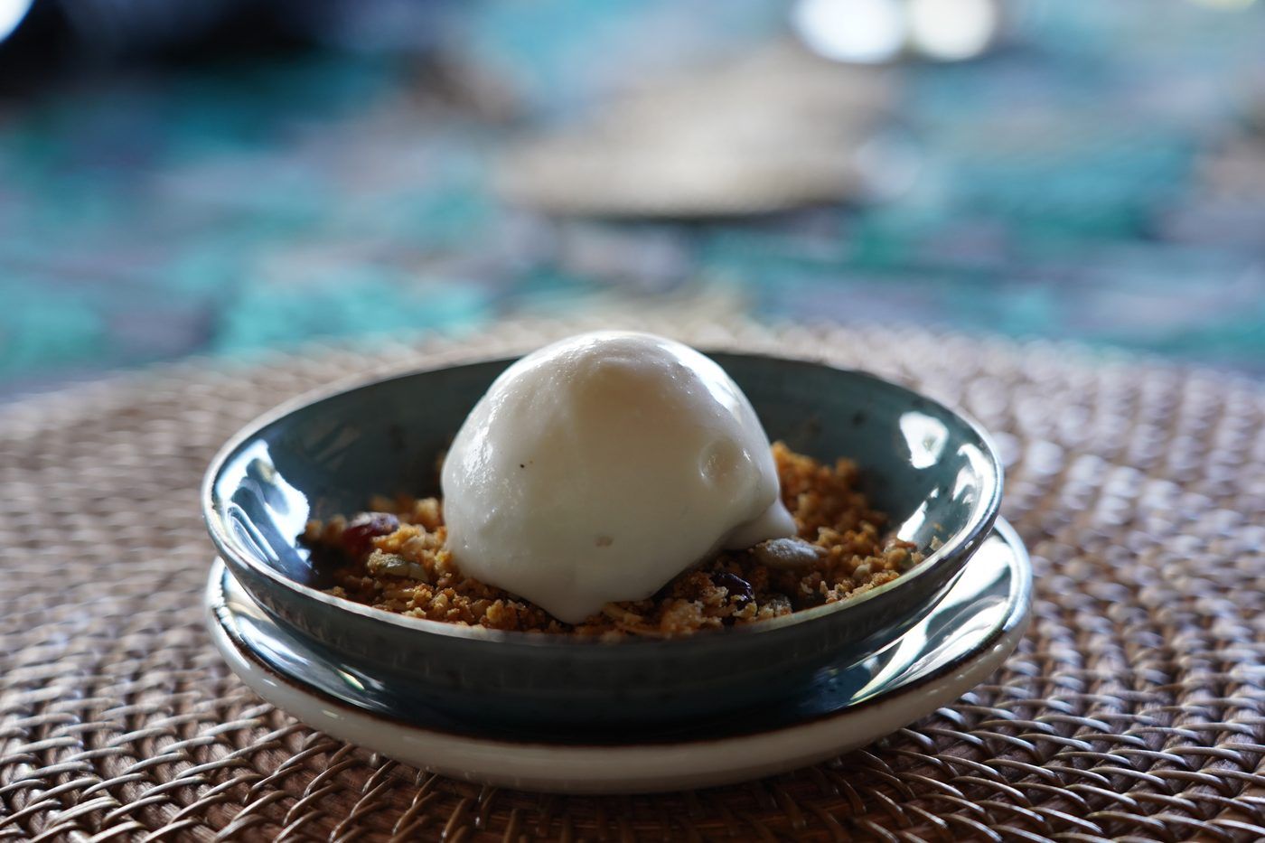 A scoop of sorbet on seeded granola crumble in a green-glazed dish set on a rattan charger.
