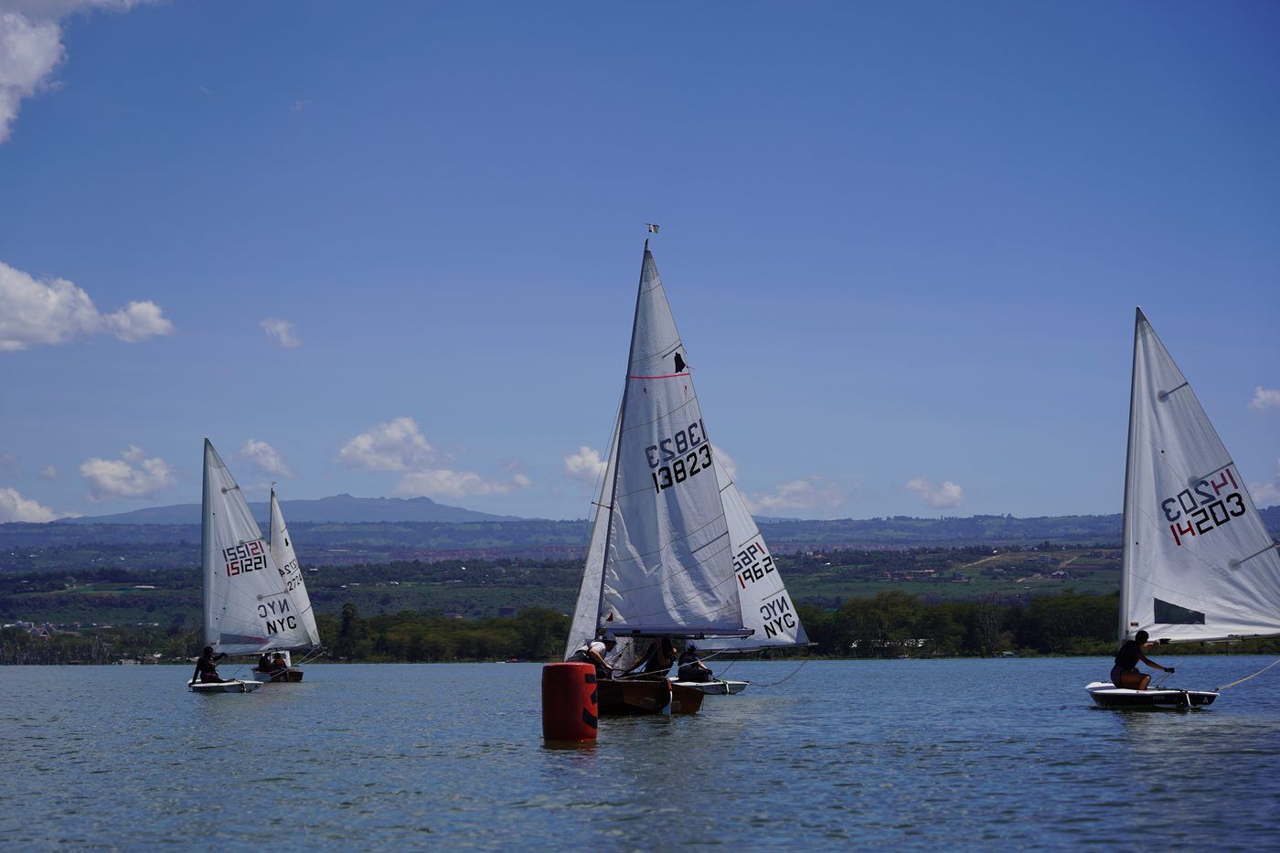 Three sailing dinghies rounding a red buoy on Lake Naivasha under a blue sky, the far shore and hazy hills beyond.