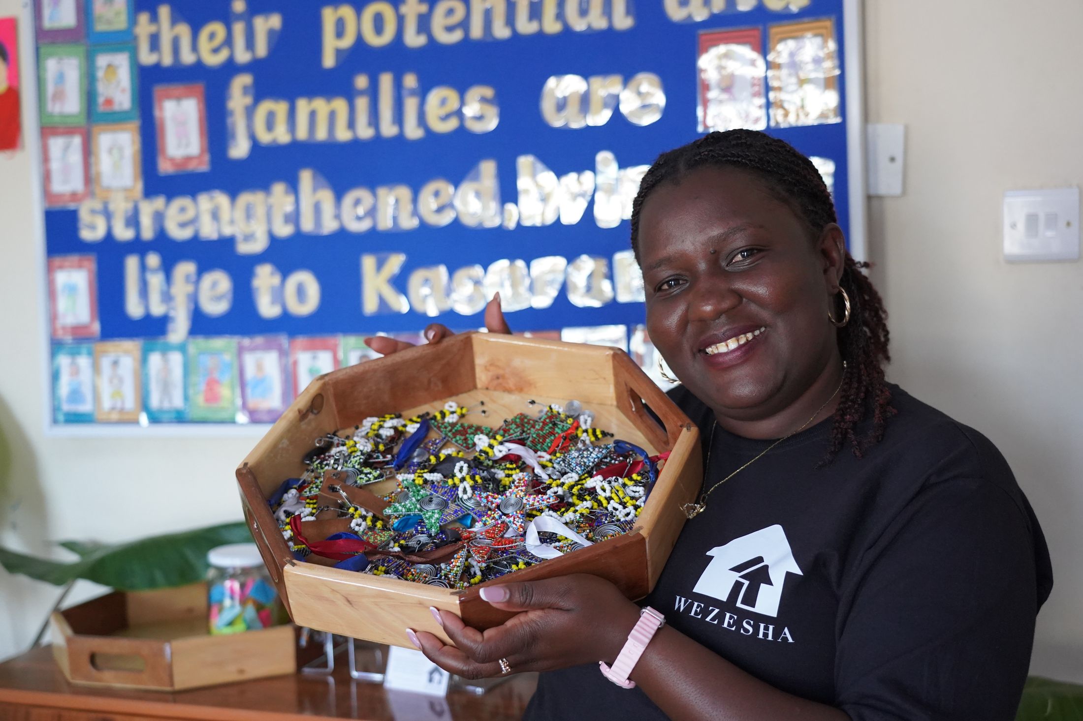 A smiling Wezesha team member holding a wooden tray filled with colourful beadwork, the centre's hand-lettered mission board behind her.