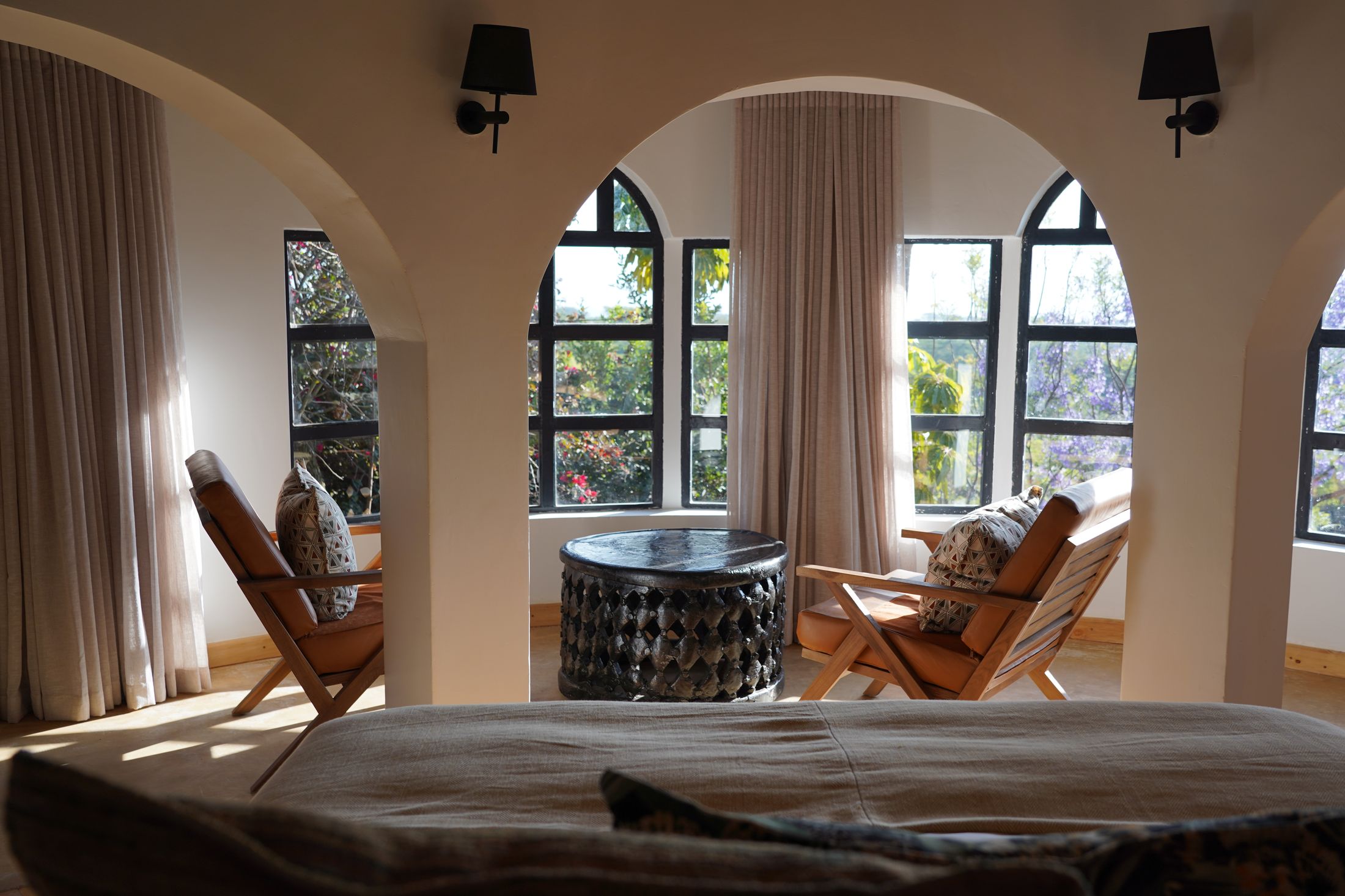 View from the bed through triple arches to leather safari chairs and a carved Bamileke table, jacaranda blooming through the windows.