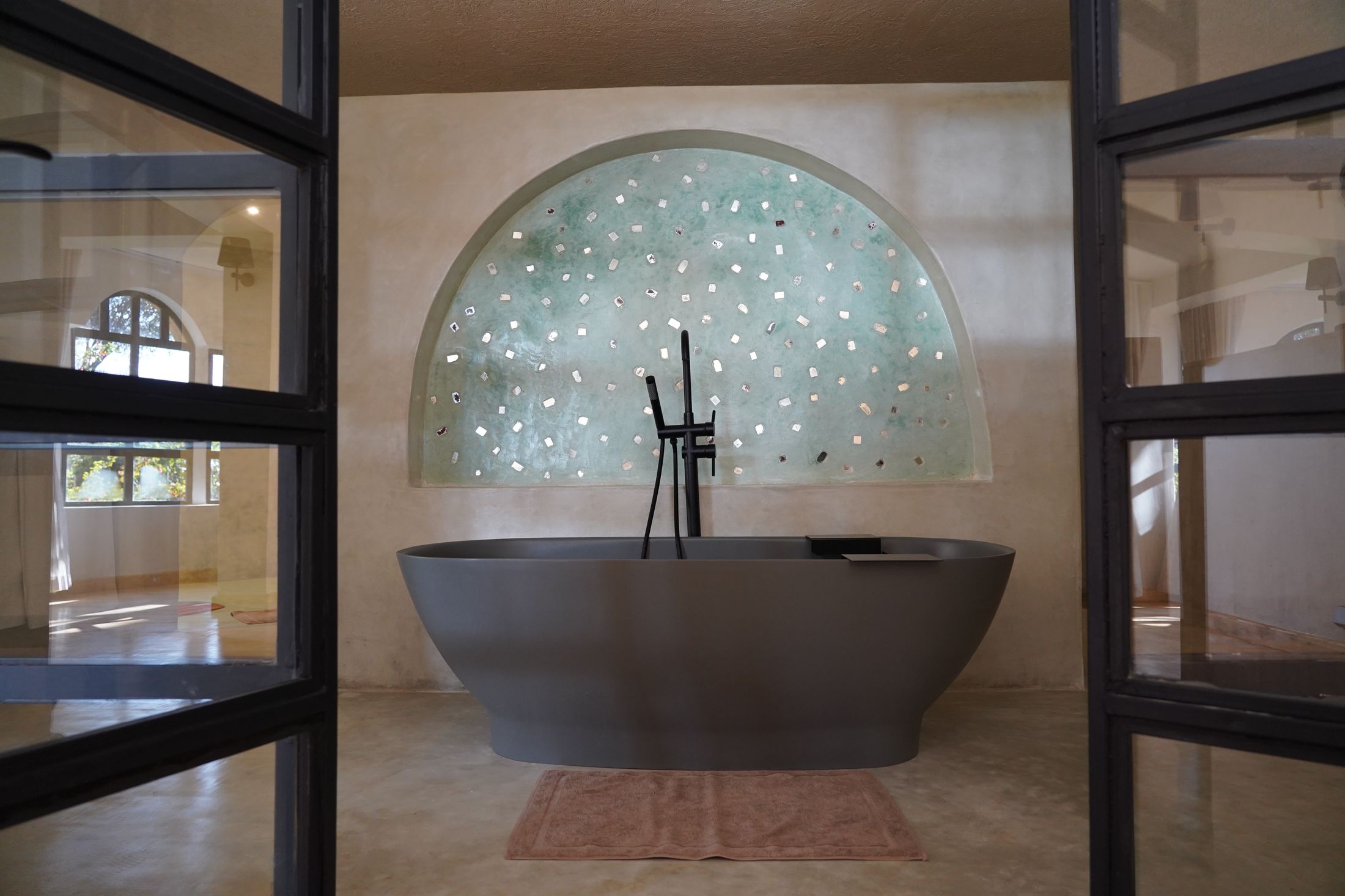 A grey stone freestanding tub centred on a sea-green plaster arch flecked with mirror, framed by black steel doors.