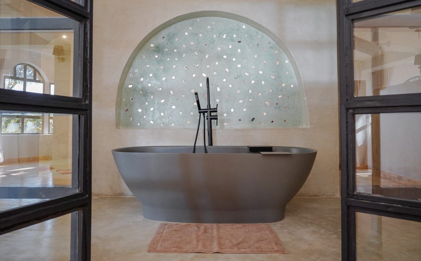 A freestanding grey stone bath centred on a half-moon plaster niche flecked with mirror, framed by black steel doors.