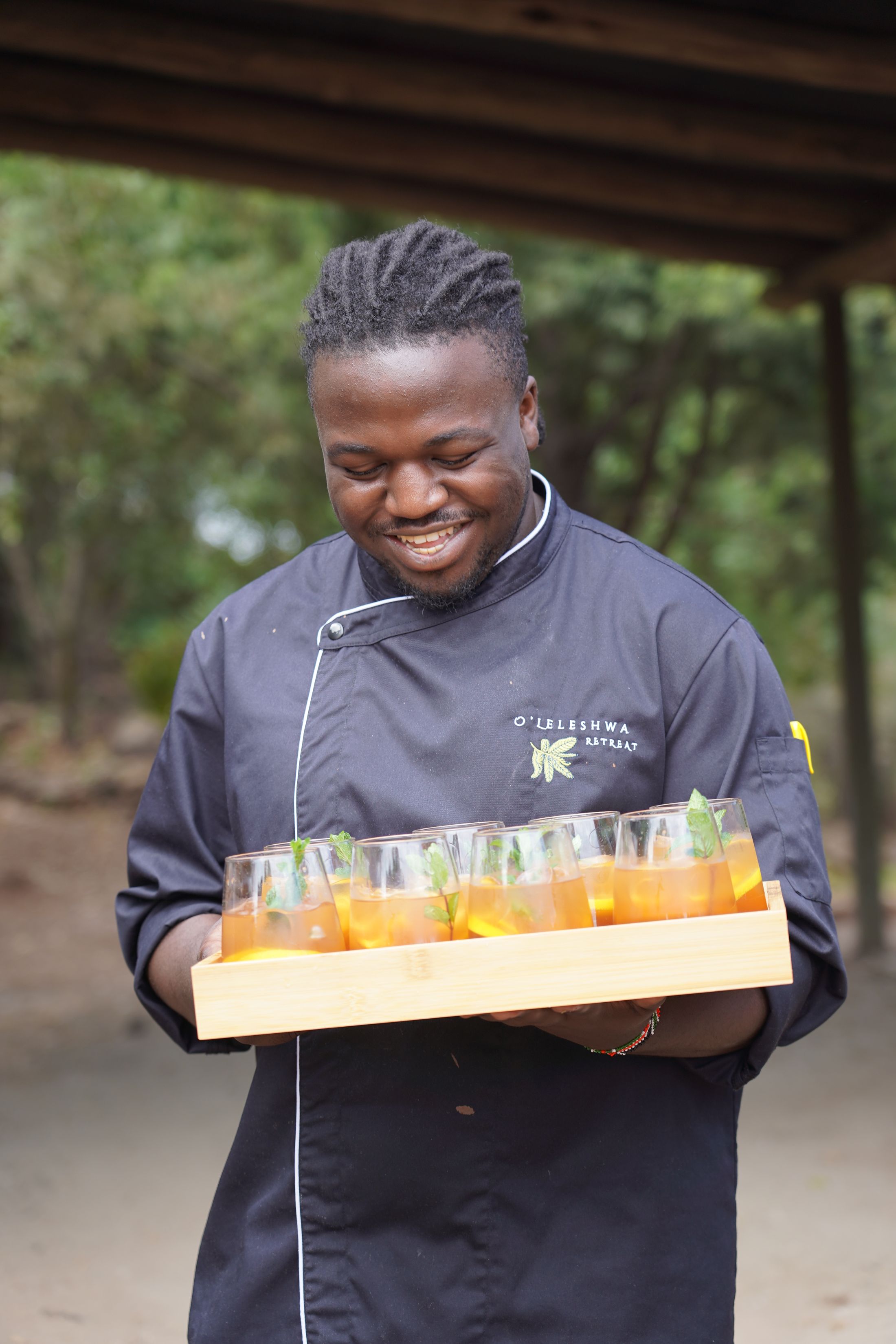 A wooden tray of iced teas with mint sprigs, carried in both hands by a chef in dark whites.
