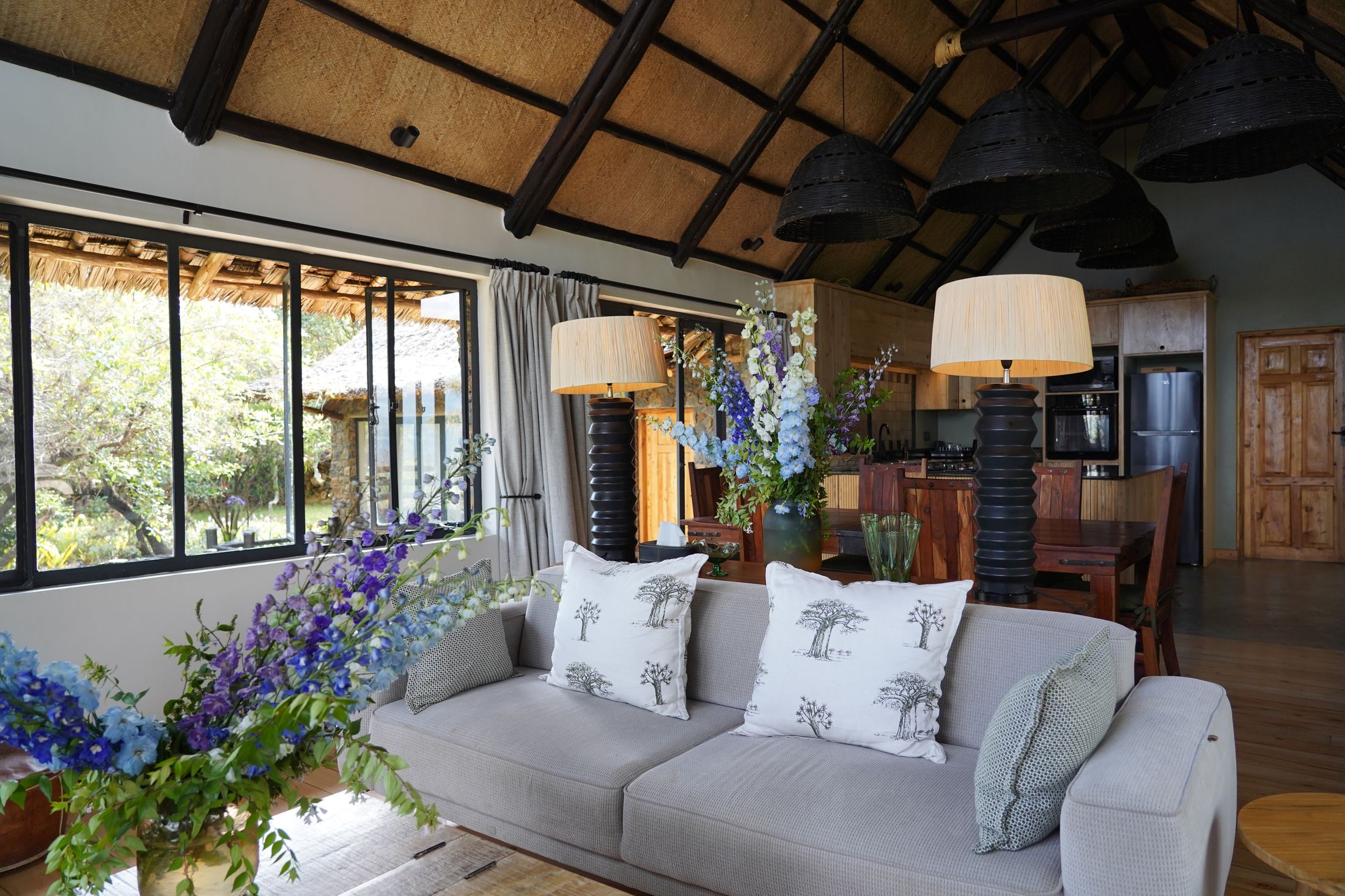 The sitting end of the great room — a grey sofa with baobab-print cushions, delphiniums on the table, the show kitchen and dining table beyond.