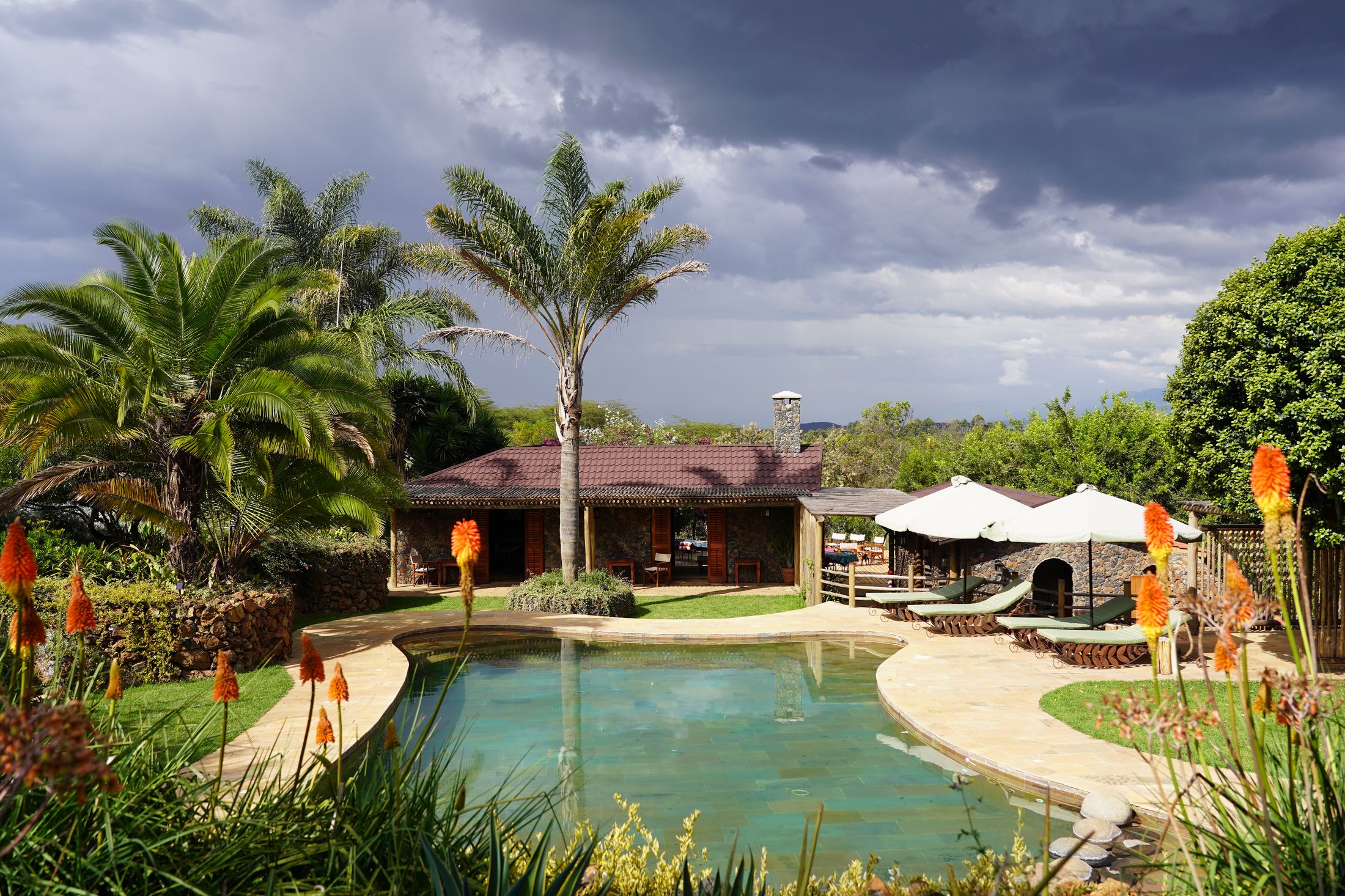 The free-form pool under a dark storm sky, red-hot poker flowers and palms in the foreground, the stone pool house with its chimney and white umbrellas on the far side.