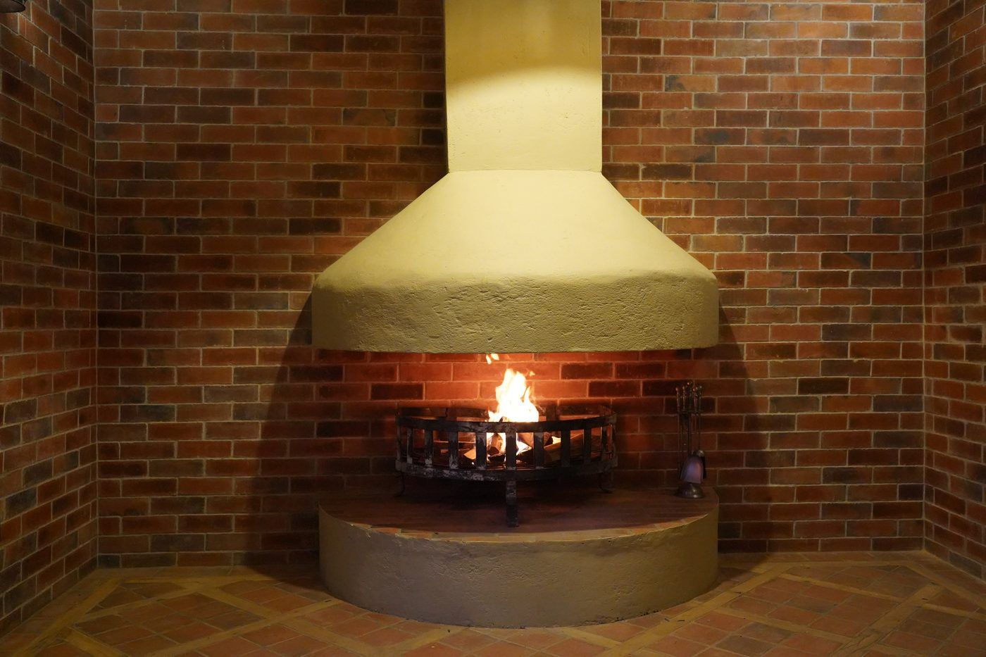 A conical plaster fireplace hood above a lit fire basket set in a brick-lined corner.