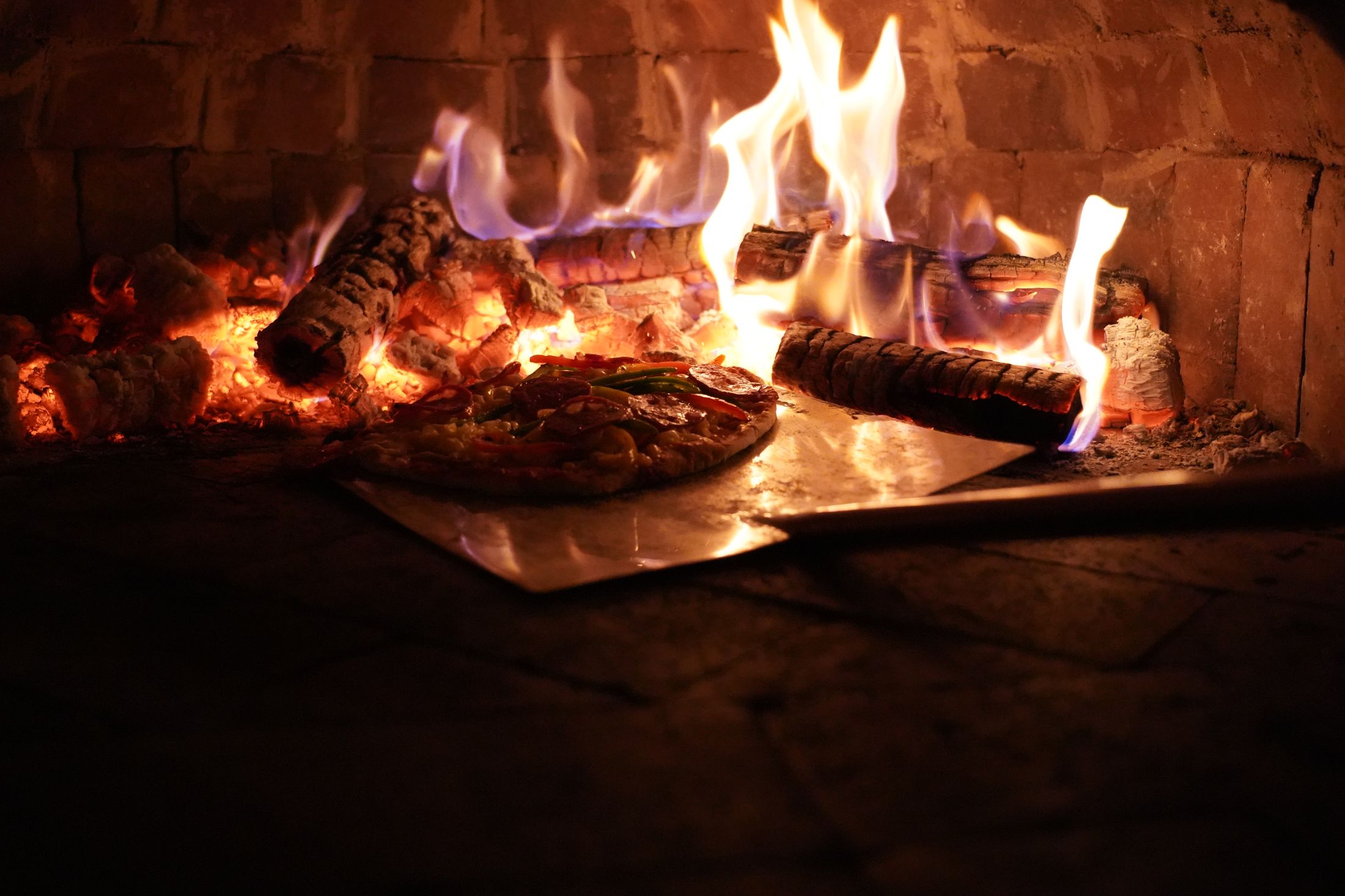 A pizza on the peel going into the estate's wood-fired brick oven, flames curling over the logs.