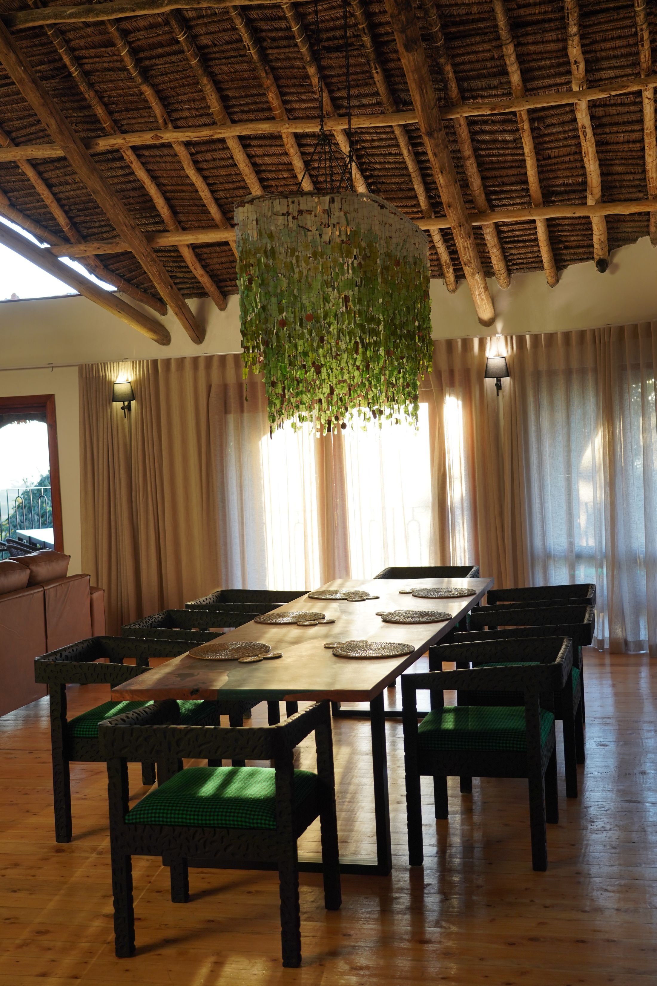 The long dining table with carved black chairs under the chandelier of repurposed Malawian glass, sun through sheer curtains.