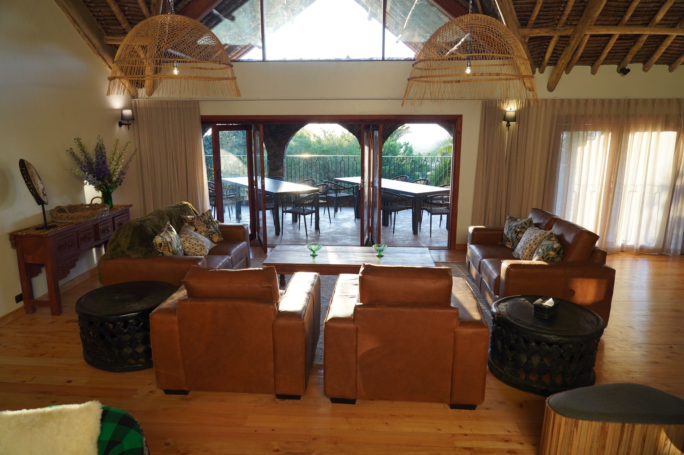The O'Leleshwa great room with its arched doors folded back — leather sofas under basket pendants, the terrace tables open to the garden beyond.