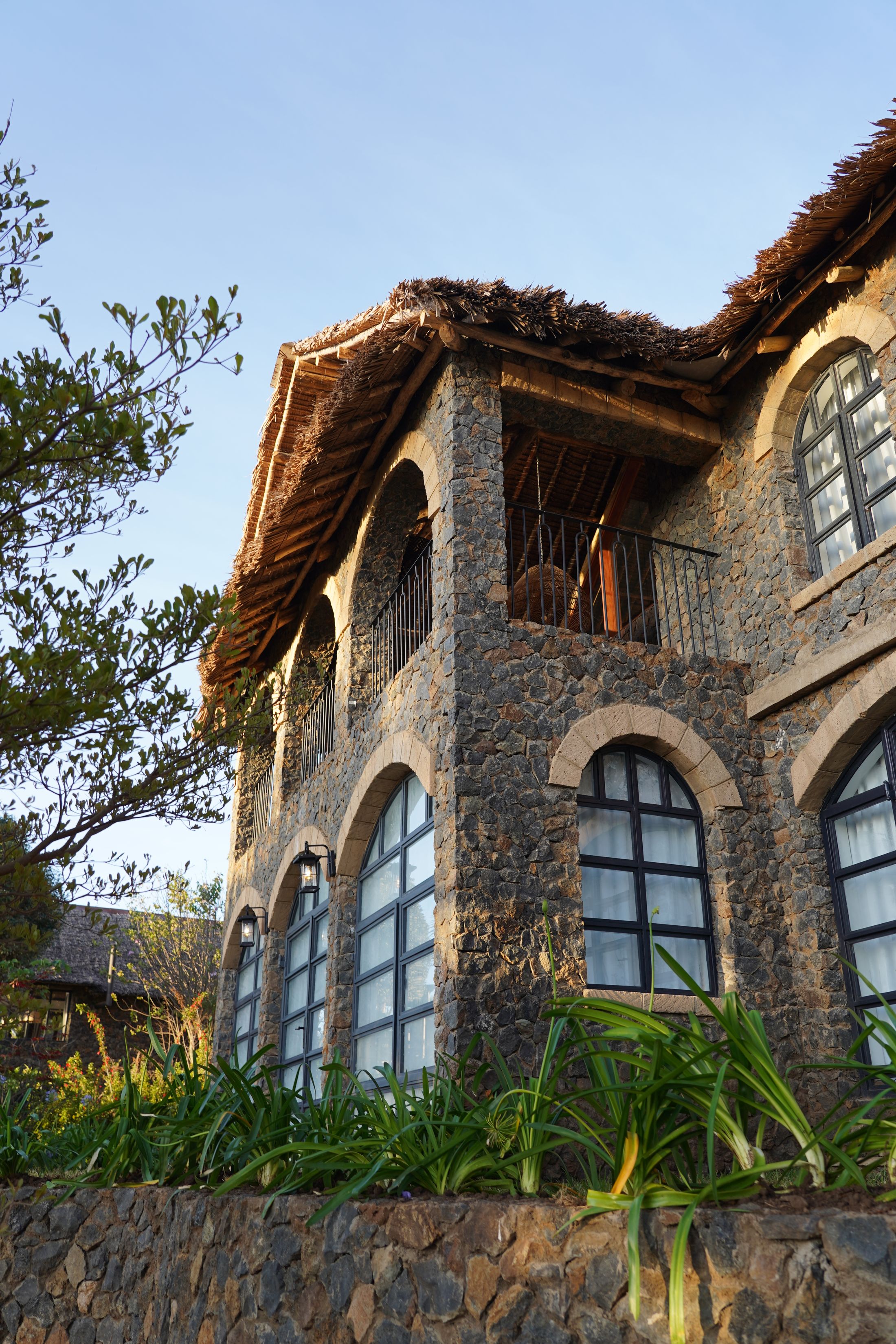 Looking up the arched stone facade and thatched balcony in golden-hour light, agapanthus on the terrace below.