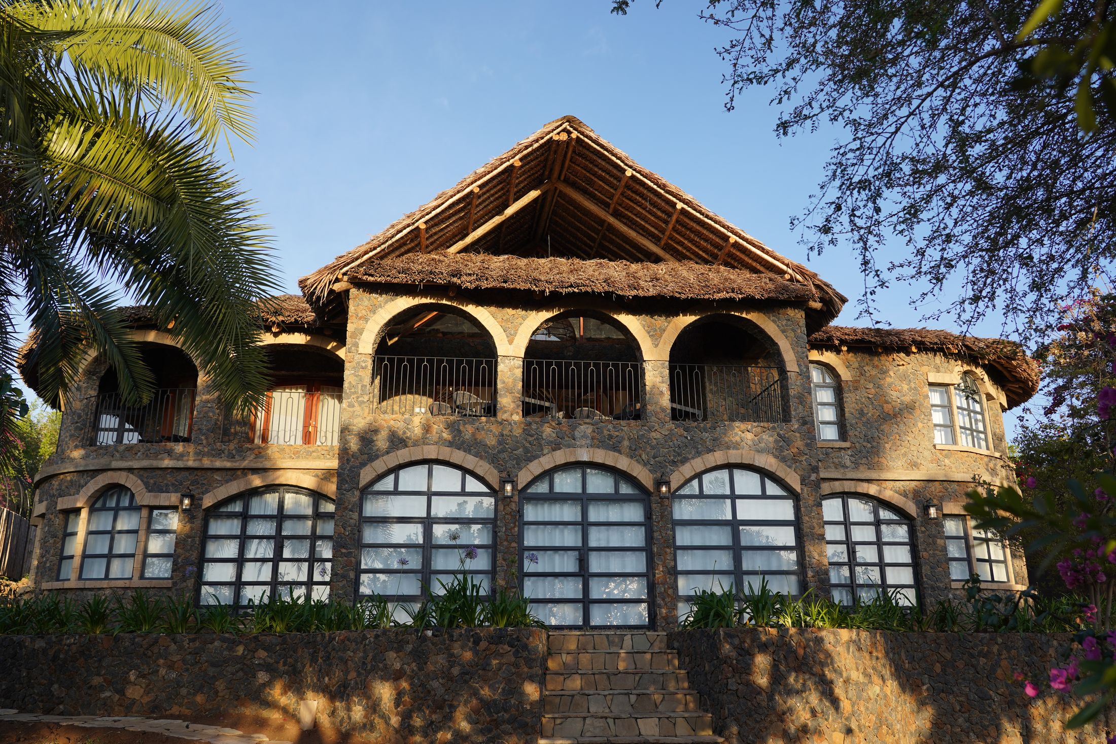 Golden-hour front elevation of the main house: stone walls, a thatched gable over a triple-arched upper loggia, arched windows below, and a dry-stone terrace wall with steps in front.