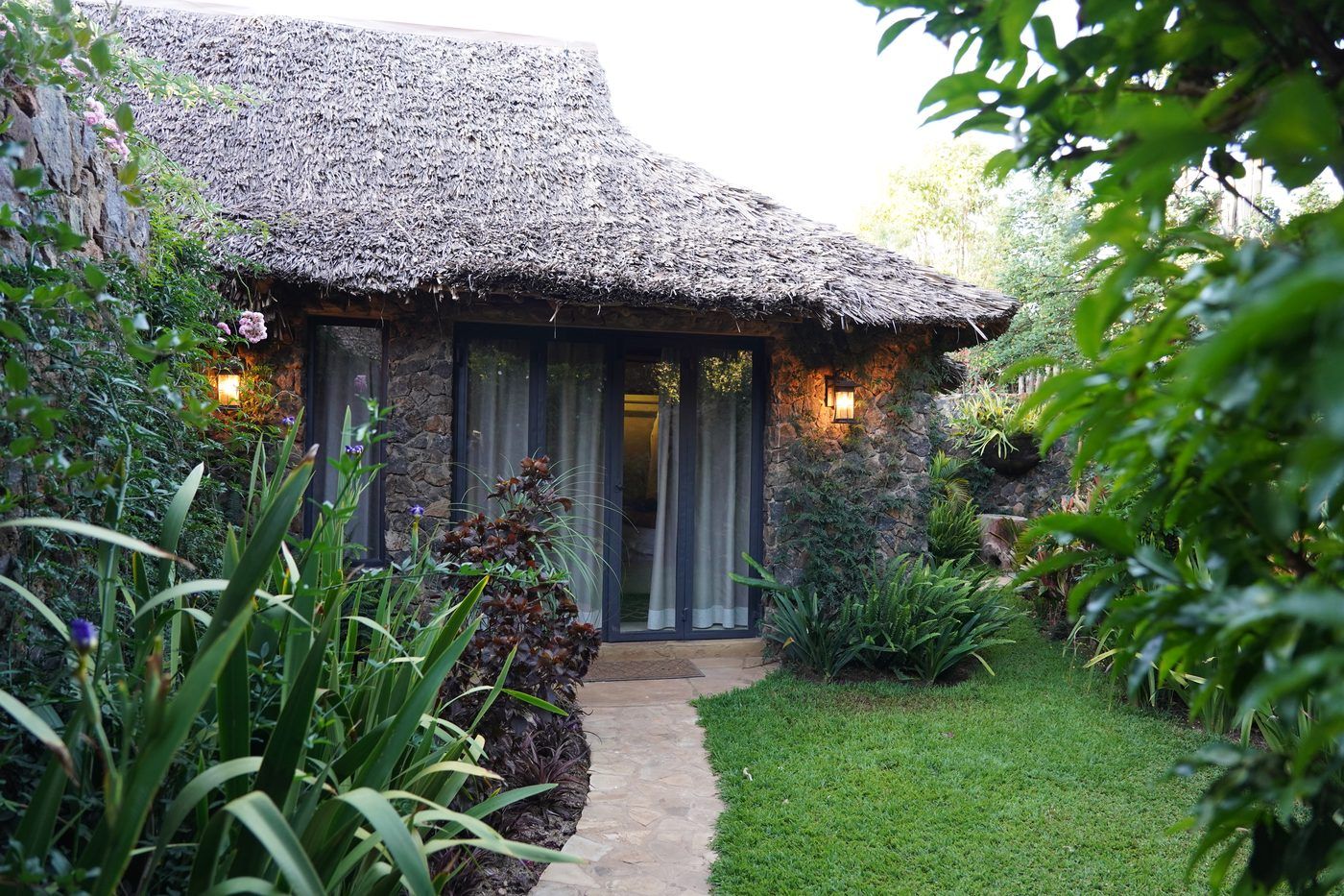 A flagstone path through planting to the glazed doors of a thatched stone garden suite, two wall lanterns lit either side of the doorway.