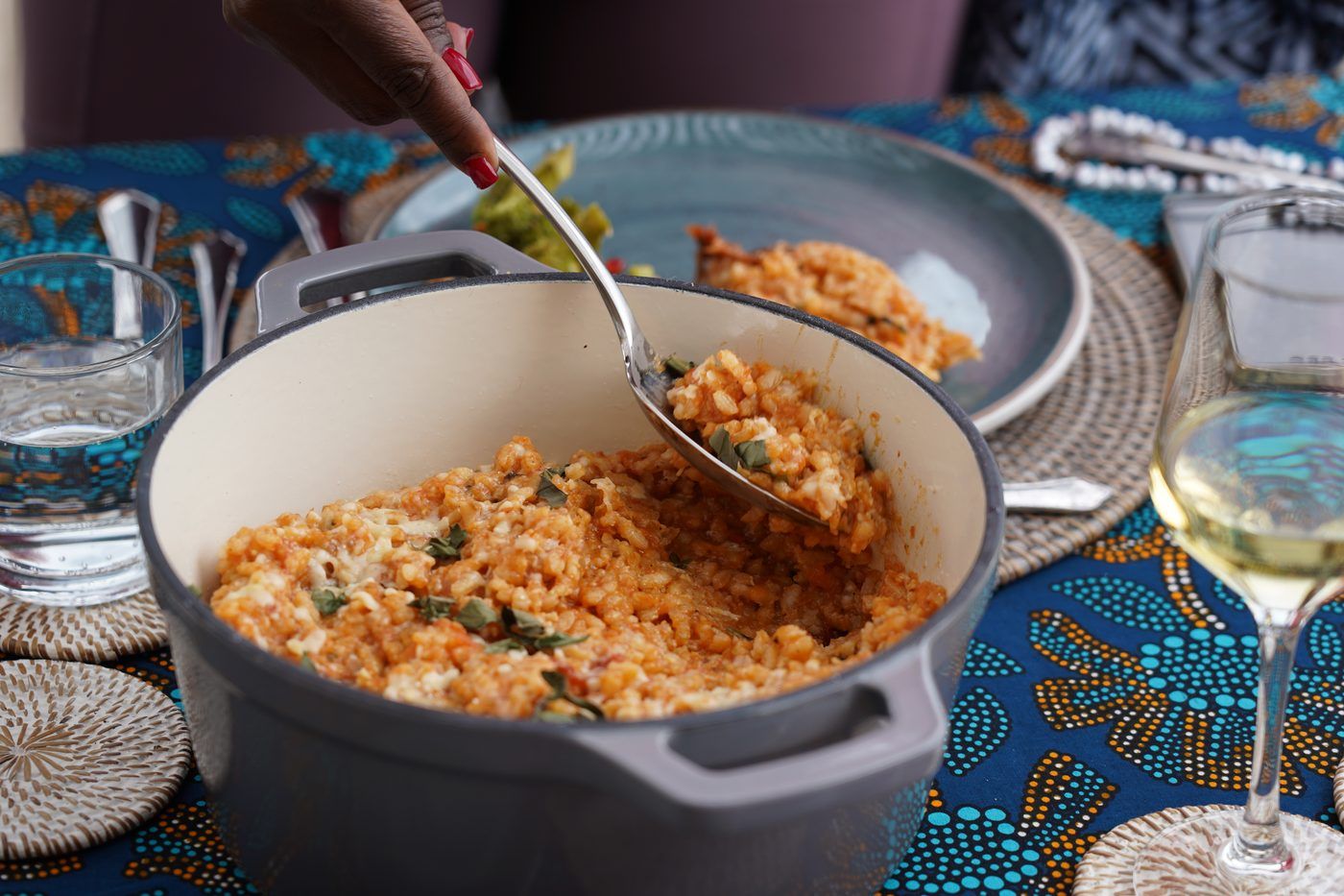 A hand spooning tomato risotto with basil from a cast-iron pot at a table dressed in blue batik, a glass of white wine beside it.