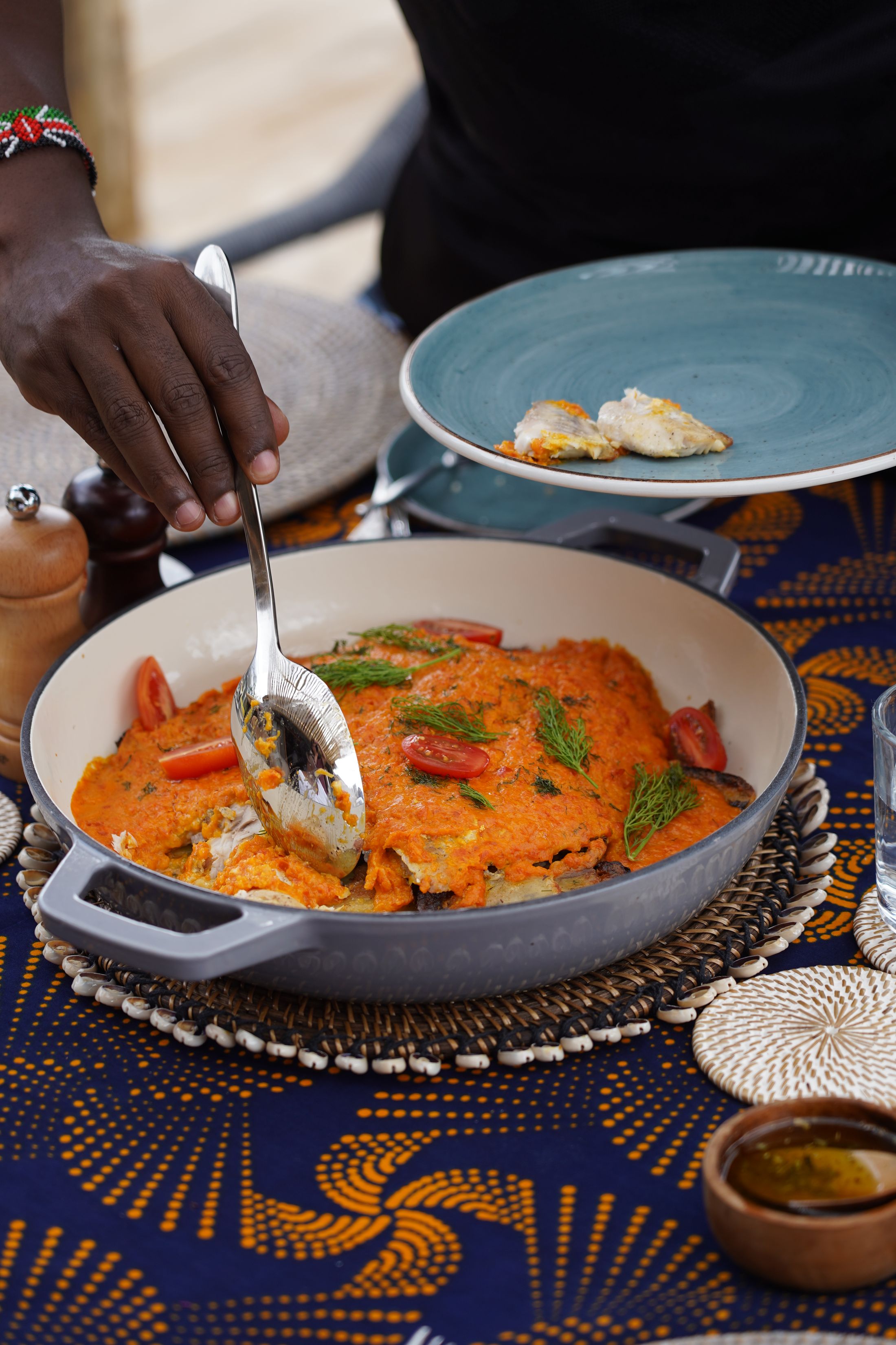 Tilapia in tomato and dill sauce being spooned from the pan at a kanga-dressed lunch table, a beaded Kenya bracelet on the server's wrist.