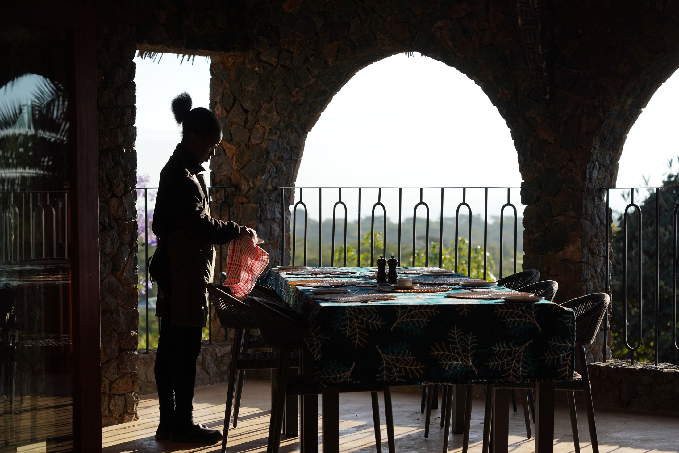 A staff member silhouetted against a stone arch, laying the breakfast table on the dining terrace in backlit morning light.