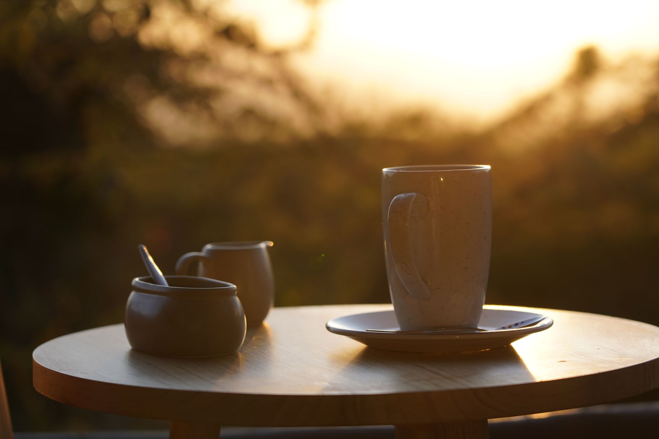 A speckled coffee mug on its saucer with sugar pot and milk jug, rim-lit by sunrise over the valley.