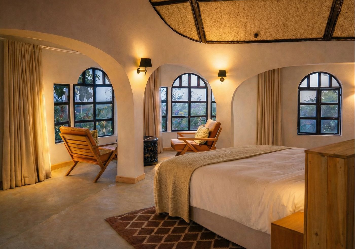 The upstairs bedroom at dusk — arched windows going blue, sconces lit, leather safari chairs in the sitting bay beyond the bed.