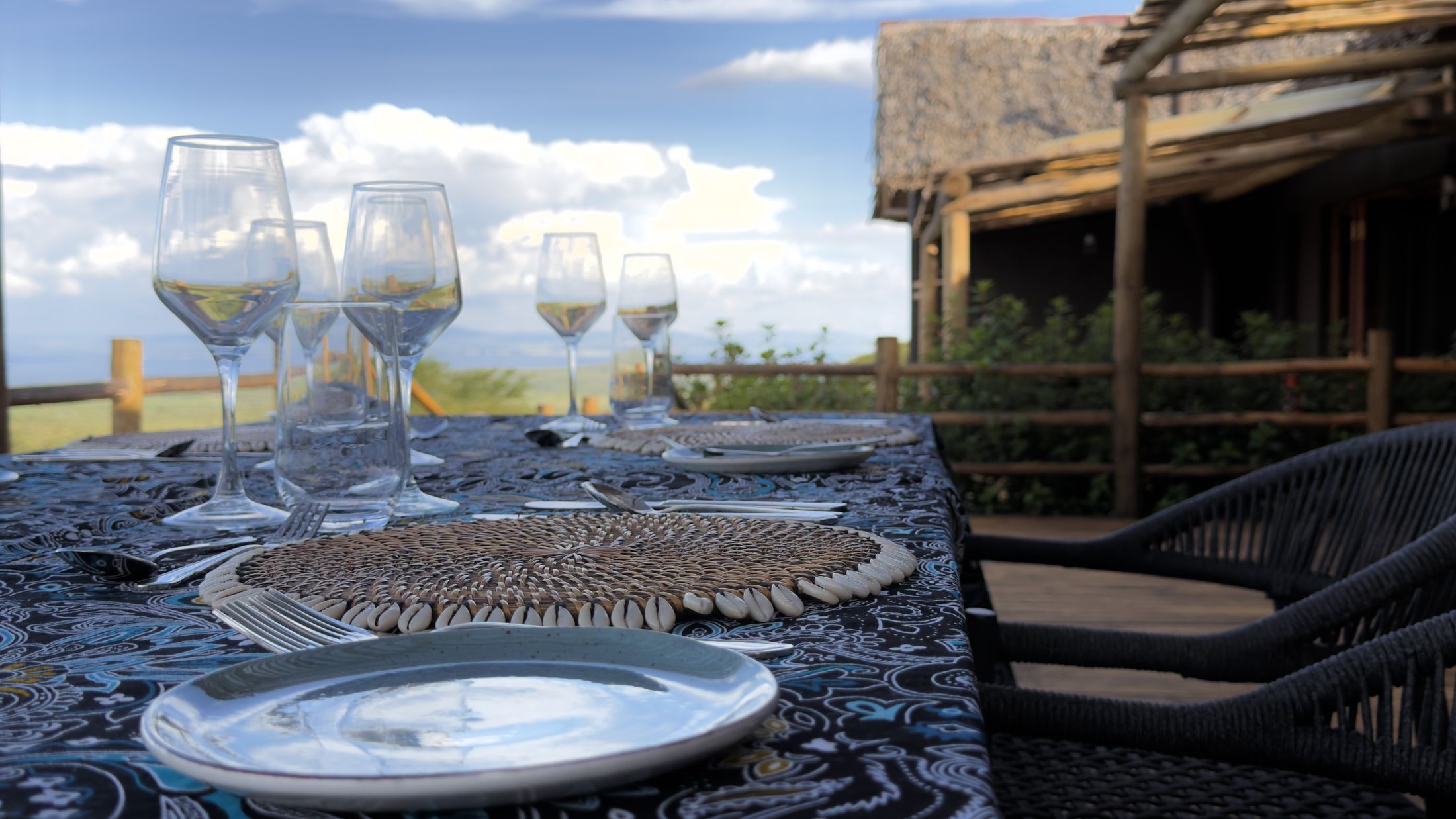 A deck table set with indigo kitenge cloth, shell-edged woven placemats and wine glasses, thatch and the valley behind.