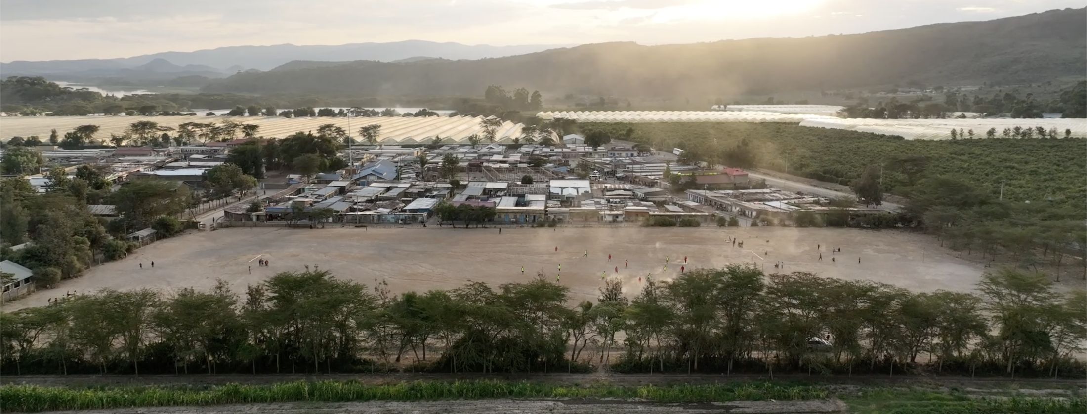 Drone view of Kasarani at dusk — a football match in progress on the dirt pitch, village rooftops and greenhouses in golden haze.