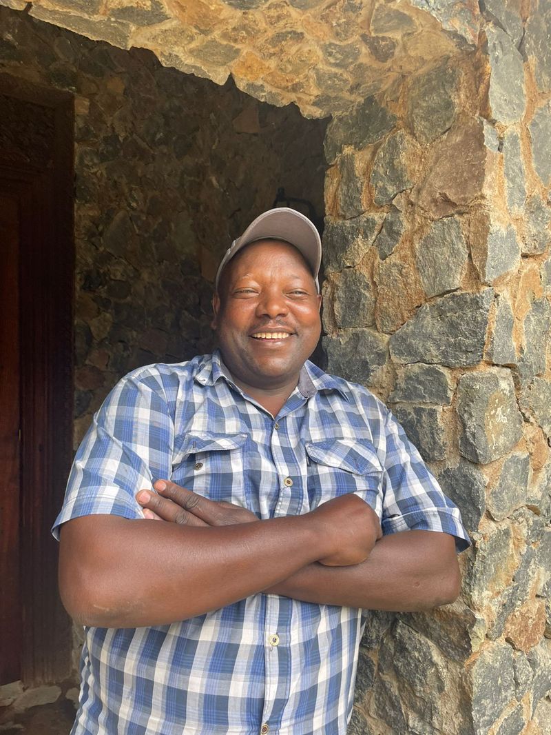 Joseph Kimani, arms crossed and smiling, leaning against the stone jamb of a doorway he built at O'Leleshwa.