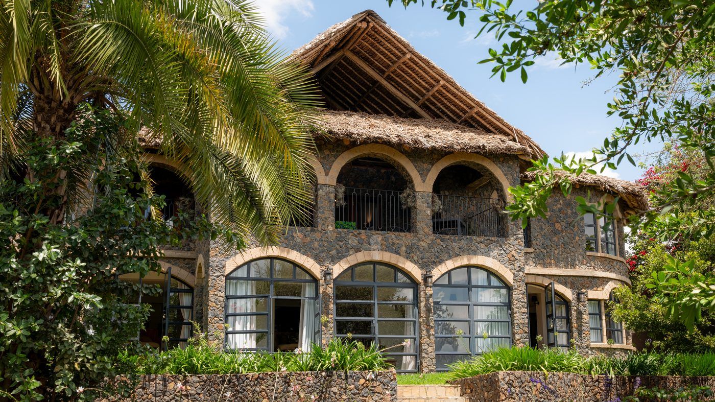 The O'Leleshwa facade seen through palm fronds: stone arches, arched steel-framed windows and a makuti thatch roof.