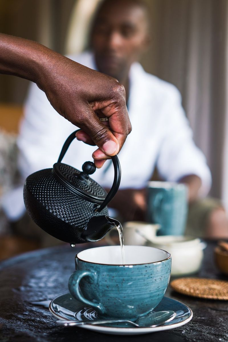 A hand pouring from a cast-iron teapot into a blue speckled cup and saucer, a figure in white soft-focused behind.