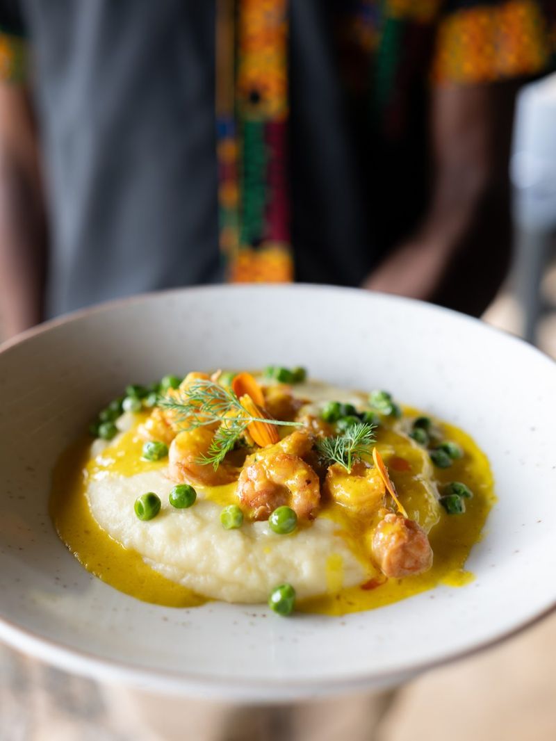 A server in a kitenge-trimmed shirt carrying a bowl of prawns on mash with peas and marigold petals.