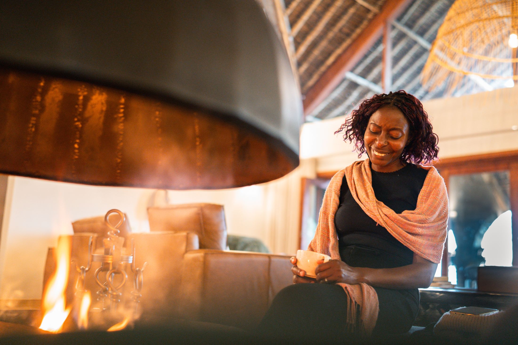 A woman in a shawl holding a bowl of tea beside the copper cone fireplace in the O'Leleshwa great room, flames at the left, thatch and a rattan pendant above.