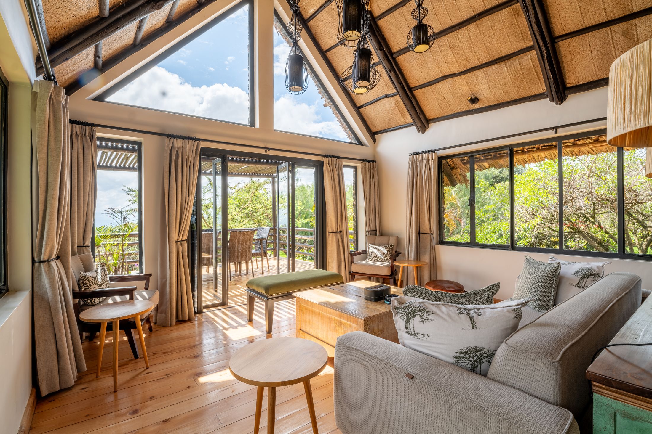The O'Lorien great room — thatch rising to a triangular skylight at the apex, dark wire-cage pendants, and glass doors open to the deck.