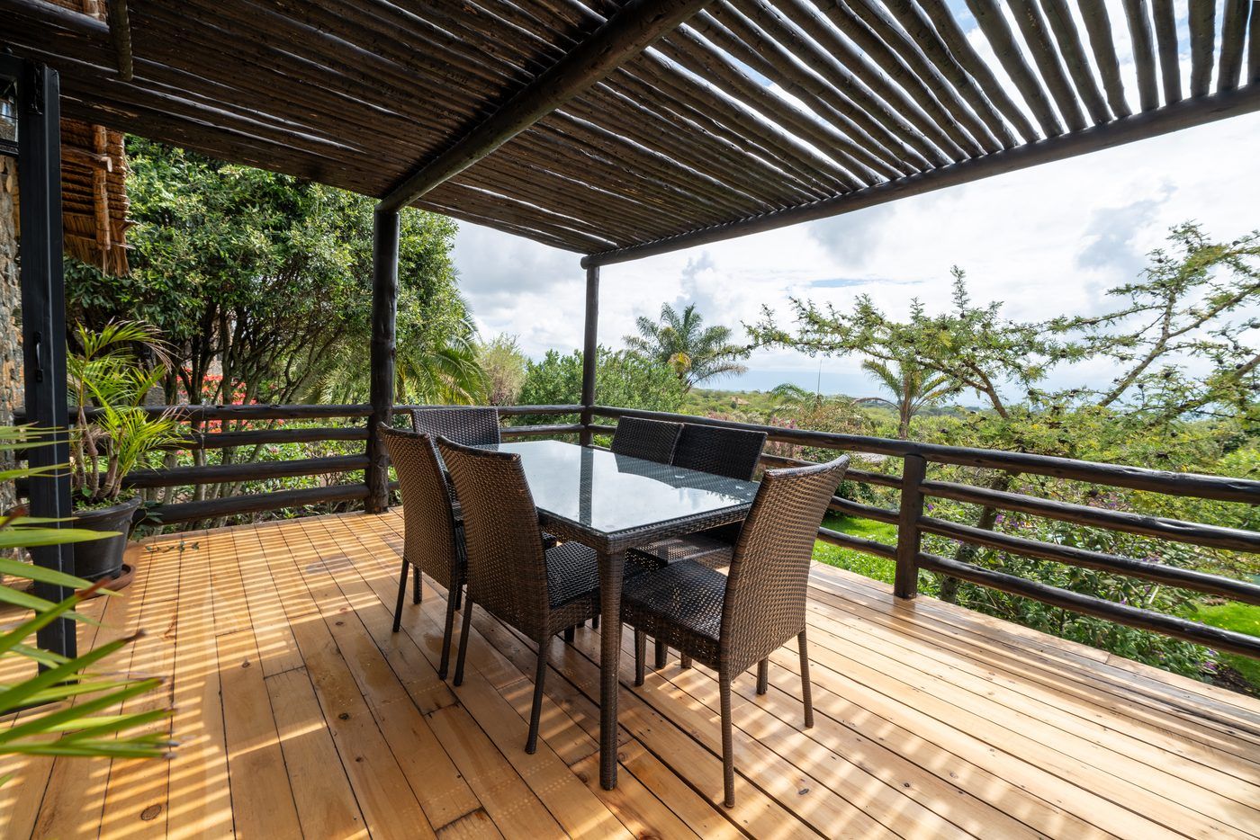 A timber deck under a pole pergola — a bare glass-topped table with six wicker chairs, acacia and bush beyond the rail, the valley opening past it.