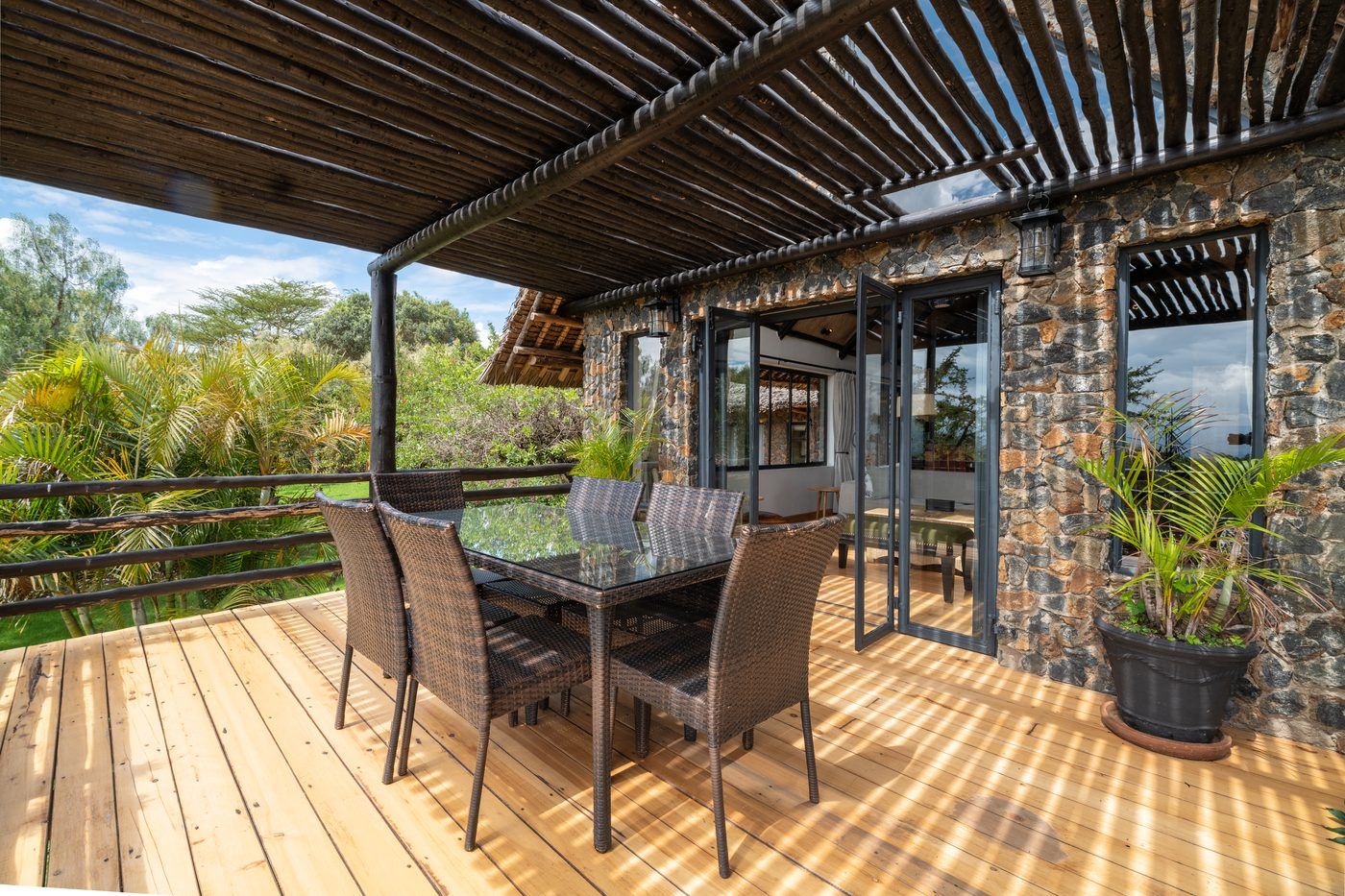 A timber deck under a pole pergola against a stone wall, a table and wicker chairs, steel bifold doors folded open to the room behind.