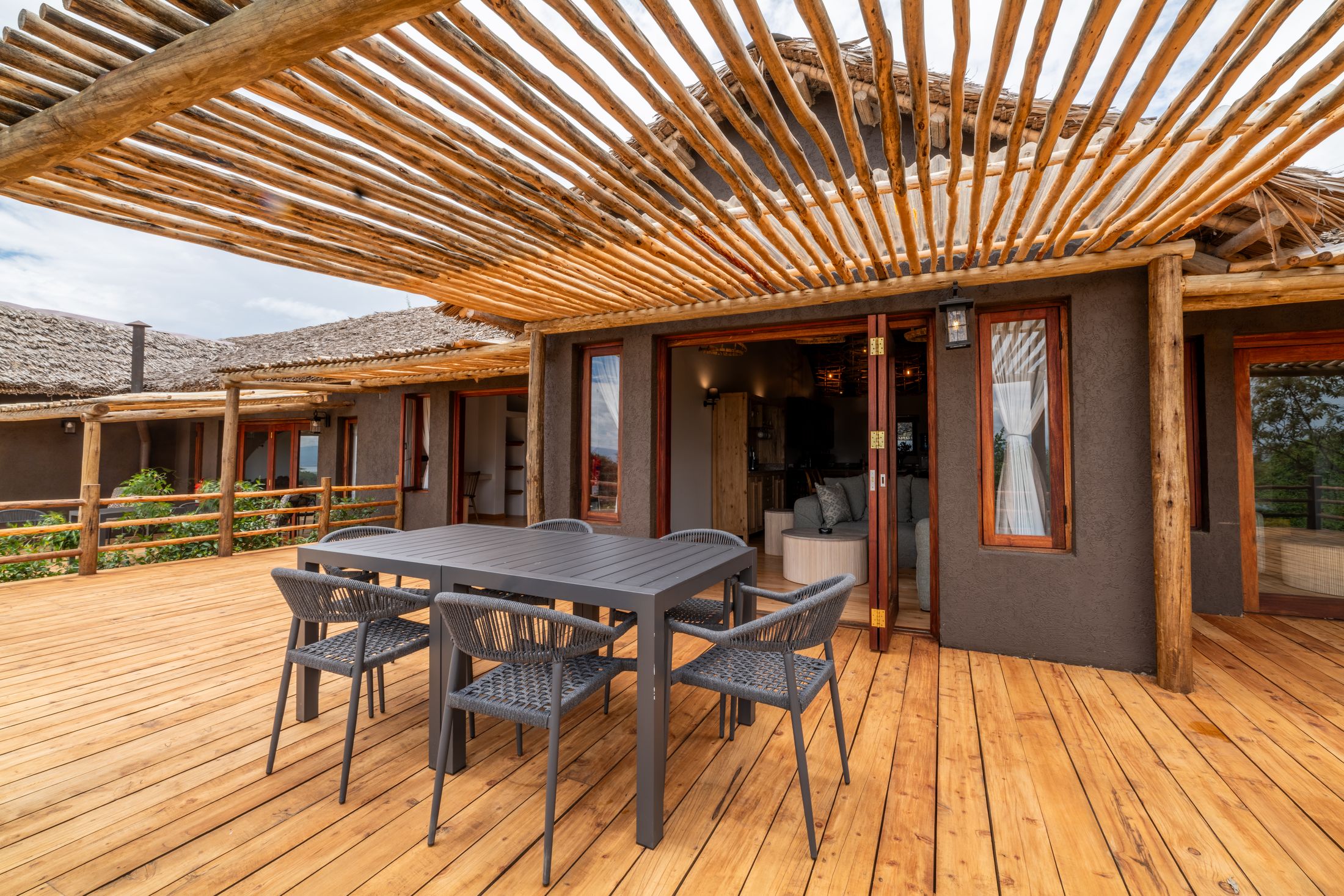 O'Lerai's timber deck under its pole pergola — a dining table and rope chairs, the folding doors open to the sitting room, thatch roofs beyond.
