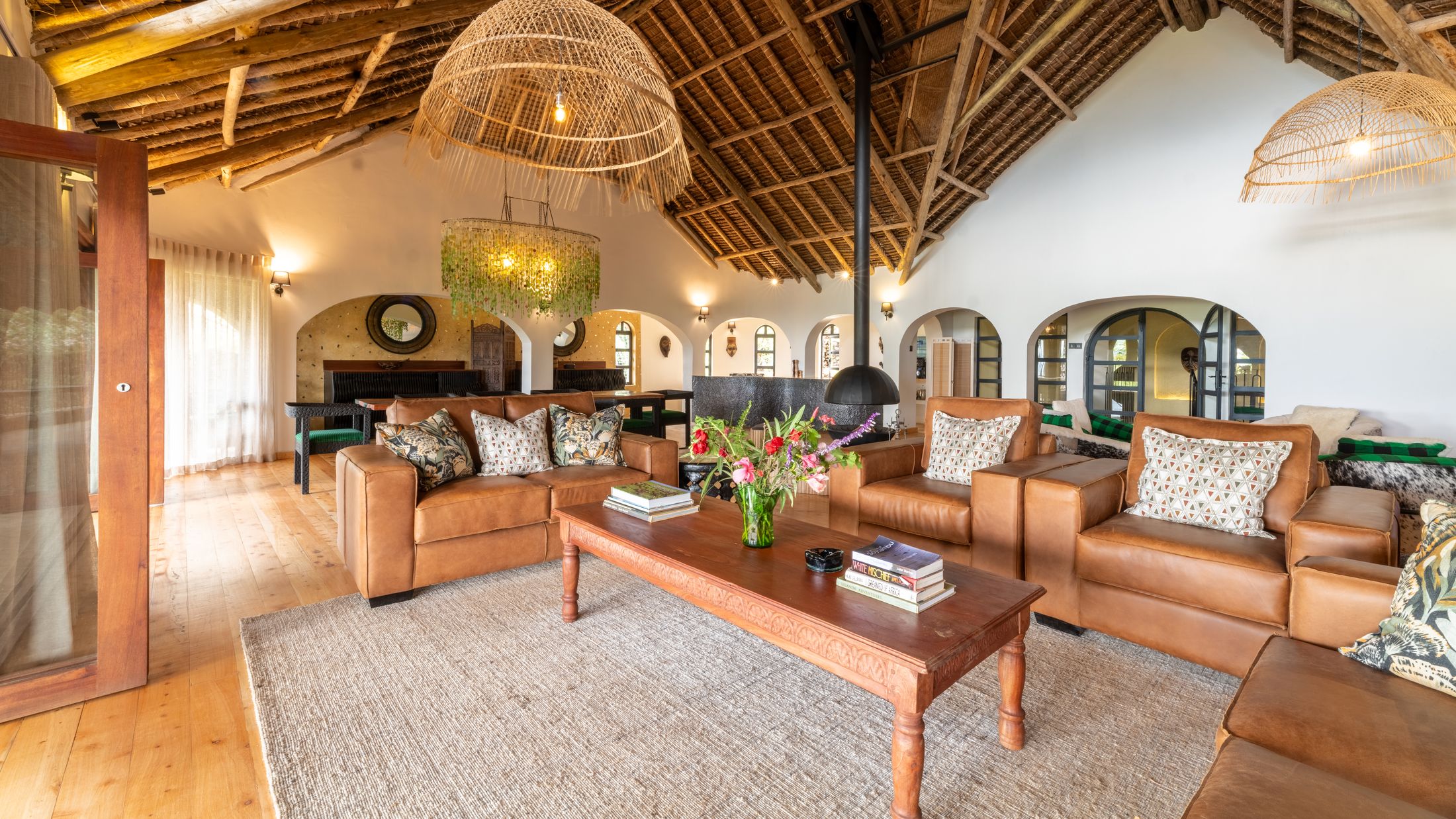 The O'Leleshwa great room under its thatch vault — leather sofas around a carved coffee table, the green Malawian-glass chandelier over the dining table beyond, arched doorways along the wall.