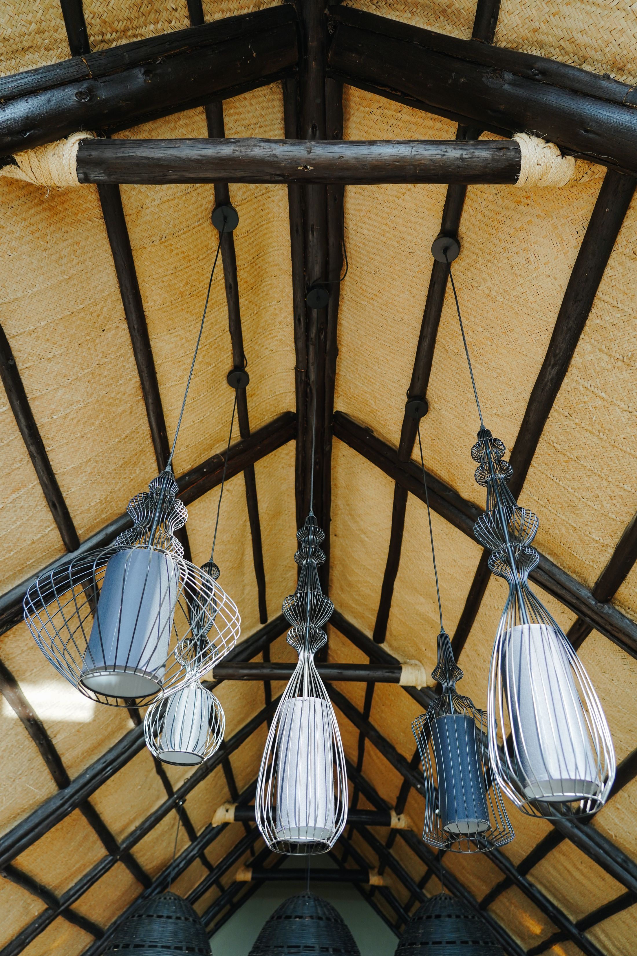 Looking straight up into the thatch: dark timber poles meeting at the ridge, a cluster of wire-cage pendant lamps hanging into the room.