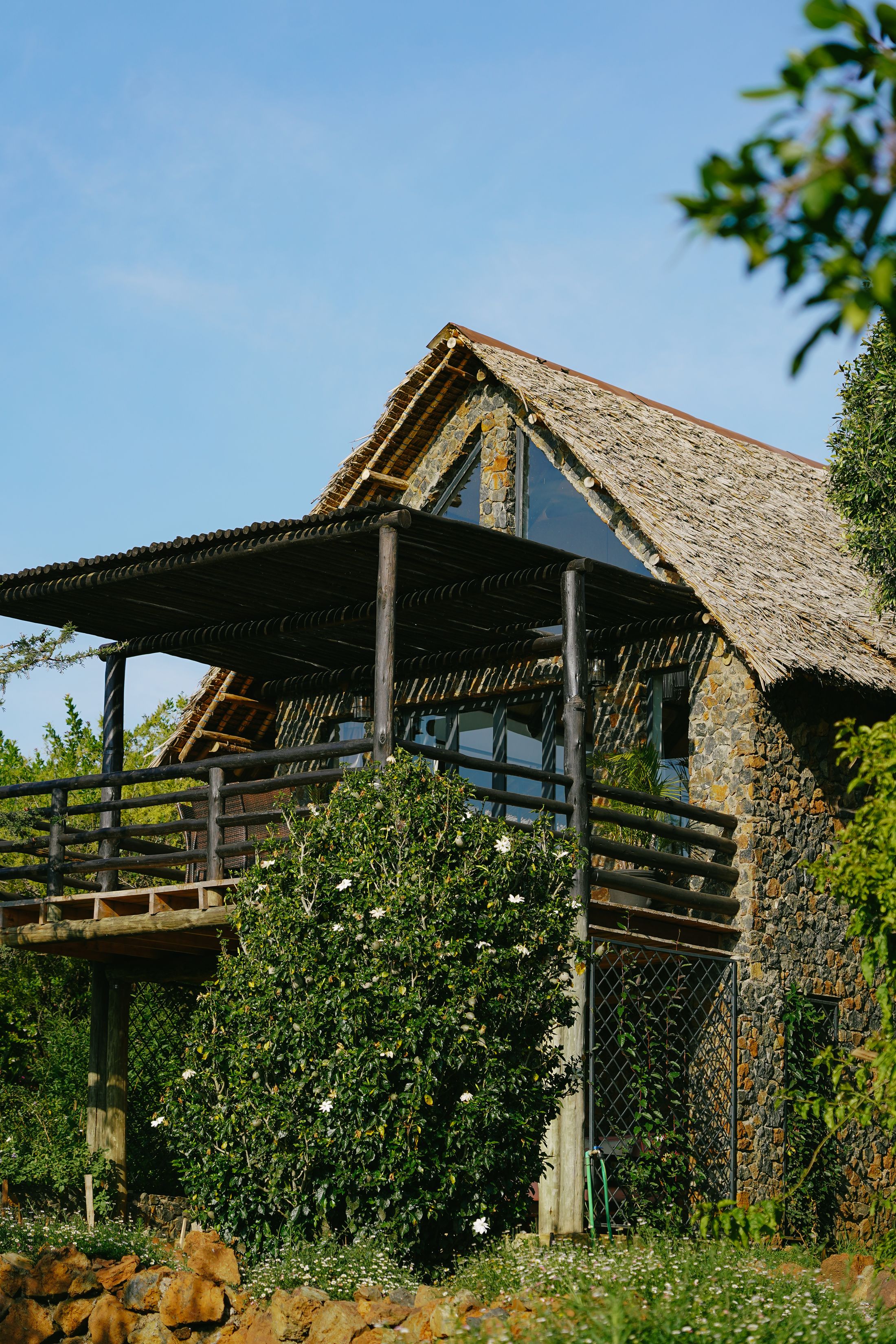 O'Lorien Villa — a steep thatched A-frame with a triangular glass gable, rising over a pole pergola deck and stone walls, blue sky above.