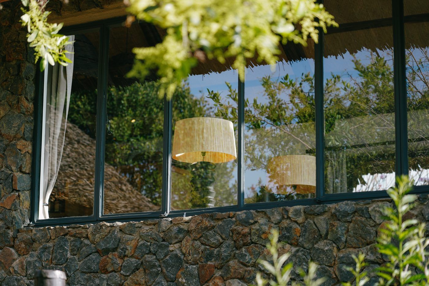 A band of steel-framed windows set into a stone wall, raffia lampshades inside and the garden reflected in the glass.