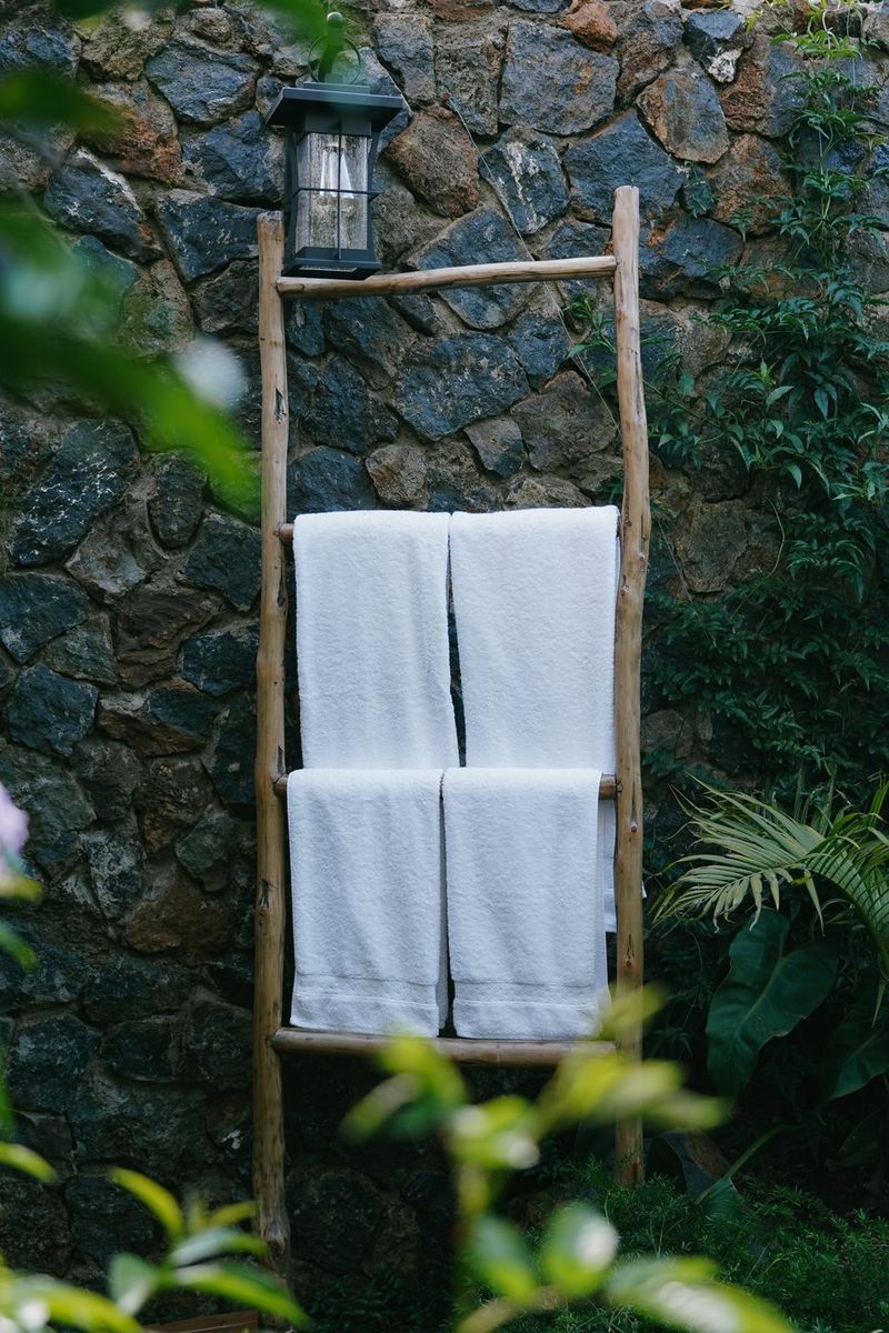 A pole towel ladder hung with white towels and topped by a lantern, against a dry-stone wall framed by garden foliage.