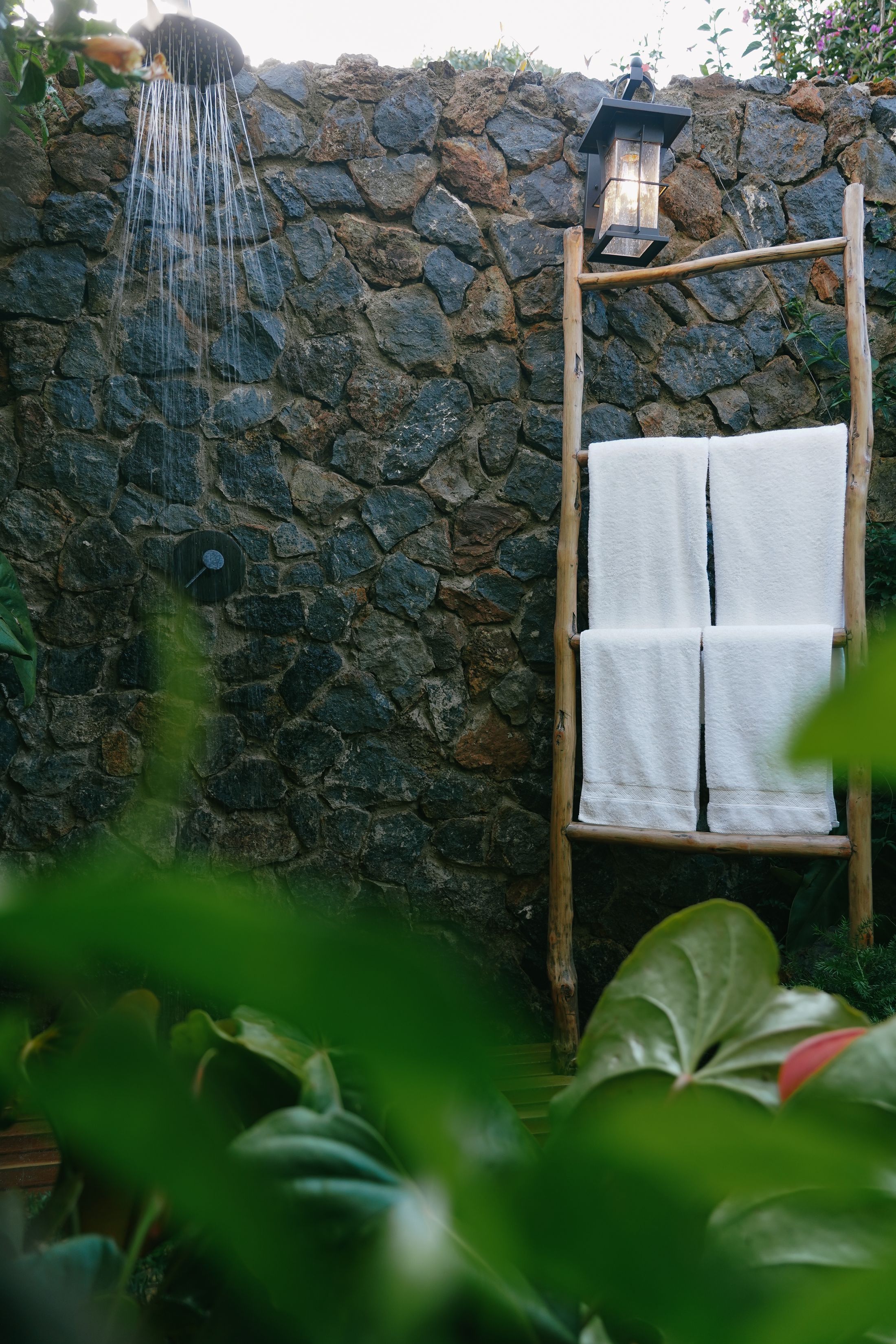 The open-air shower running against the stone wall of its court, a lantern and white towels on the pole ladder beside it, foliage in the foreground.