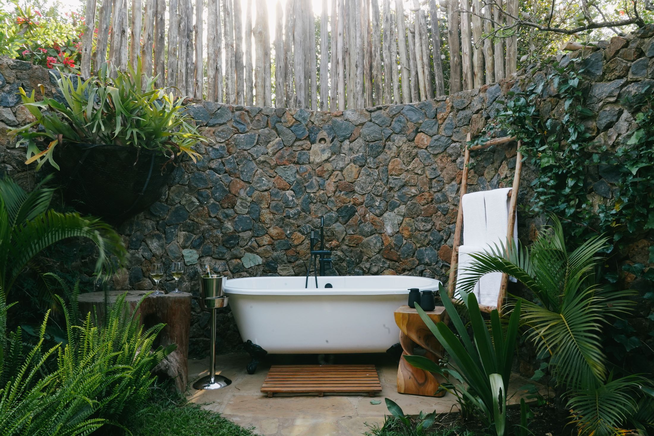 The O'Lorien bath court — a white claw-foot tub against a stone wall, ferns and a staghorn basket above, the pole fence backlit by low sun, wine cooling beside the tub.