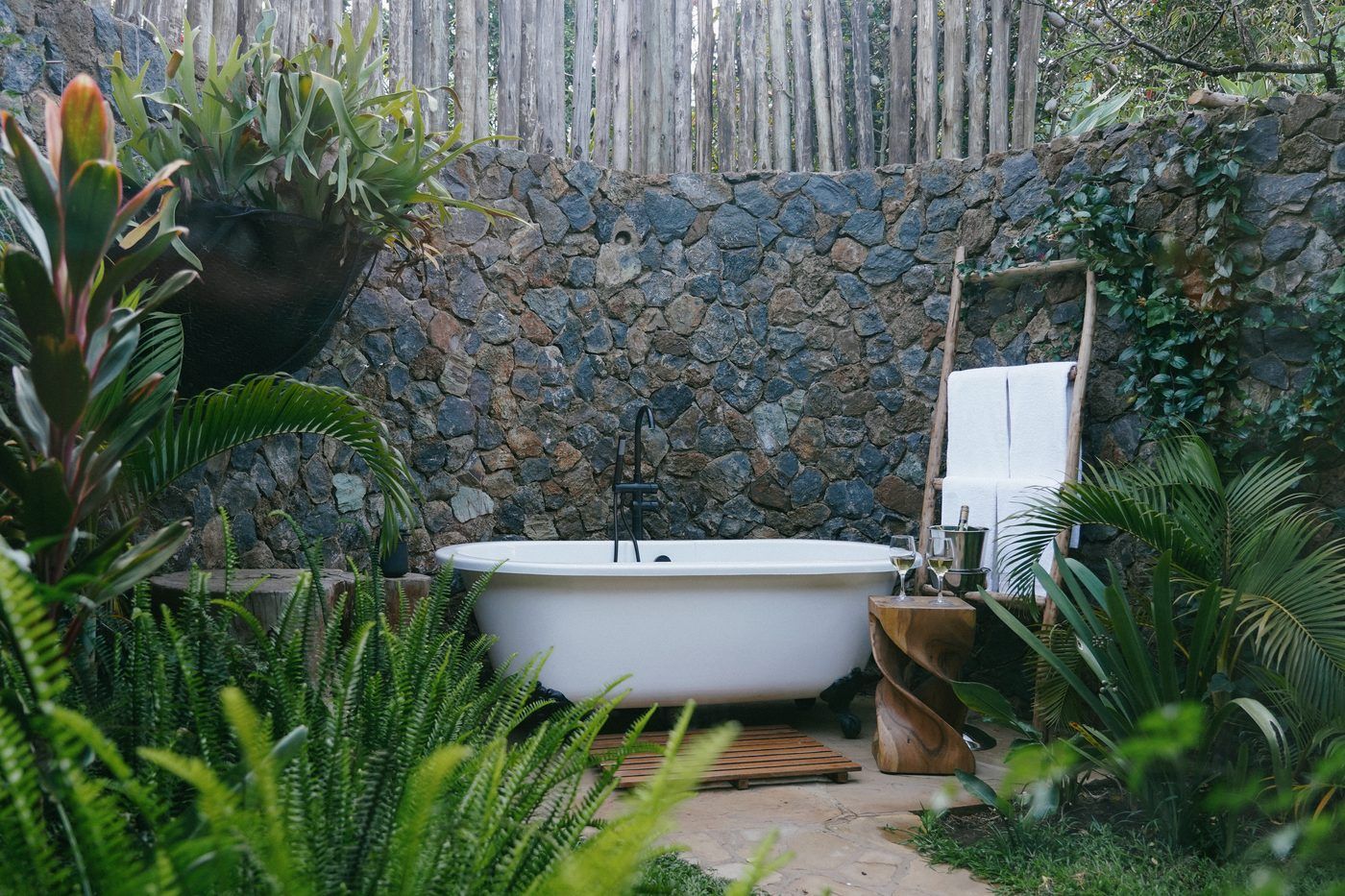 A clawfoot bath against the stone wall of O'Lorien's walled garden in soft shade, ferns and cordyline in the foreground, towels on a ladder.