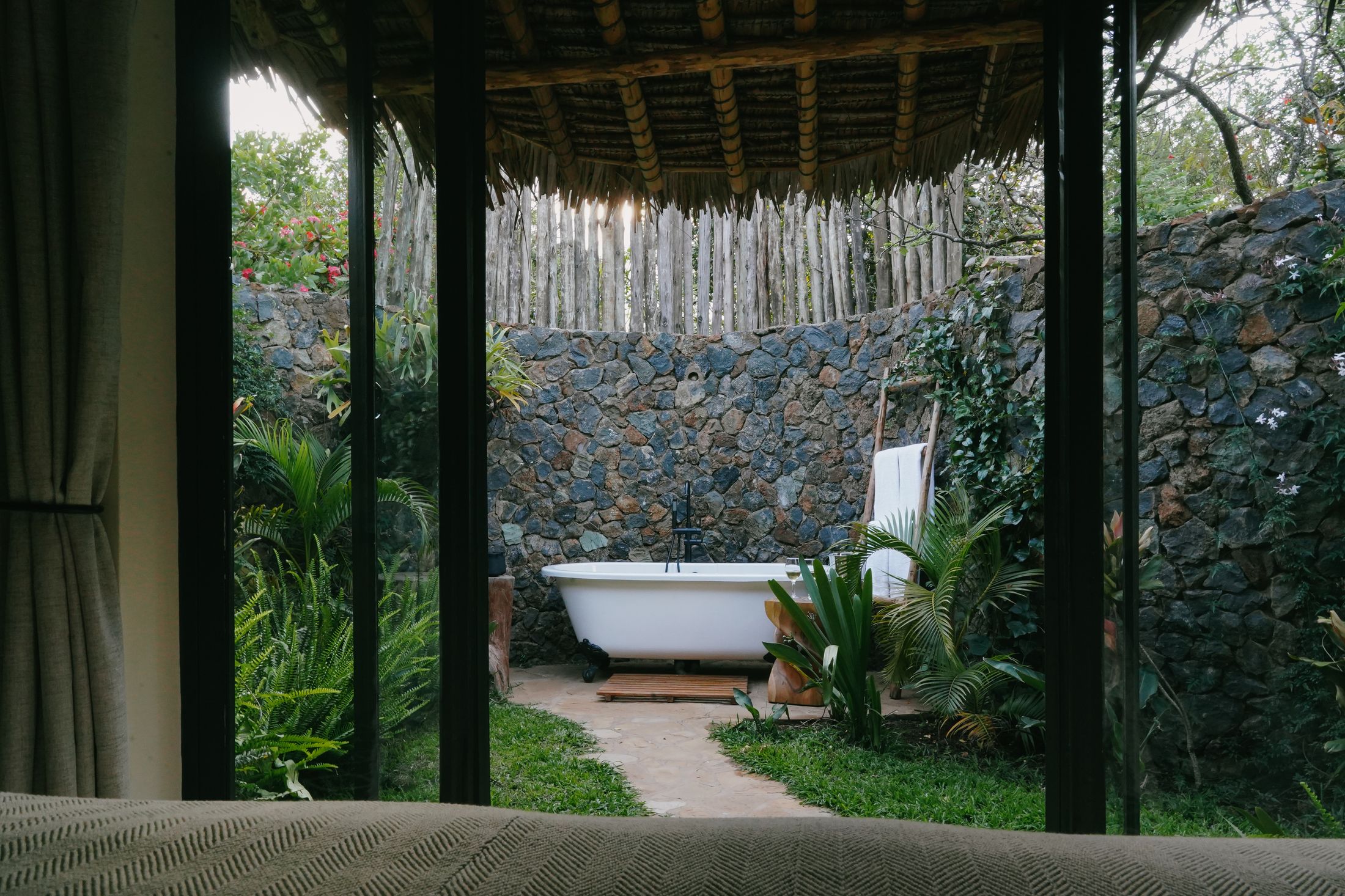 View from an O'Lorien bed through open steel-framed glass doors: a grass path leading to the claw-foot bath against the stone wall of the court, thatch eave above.