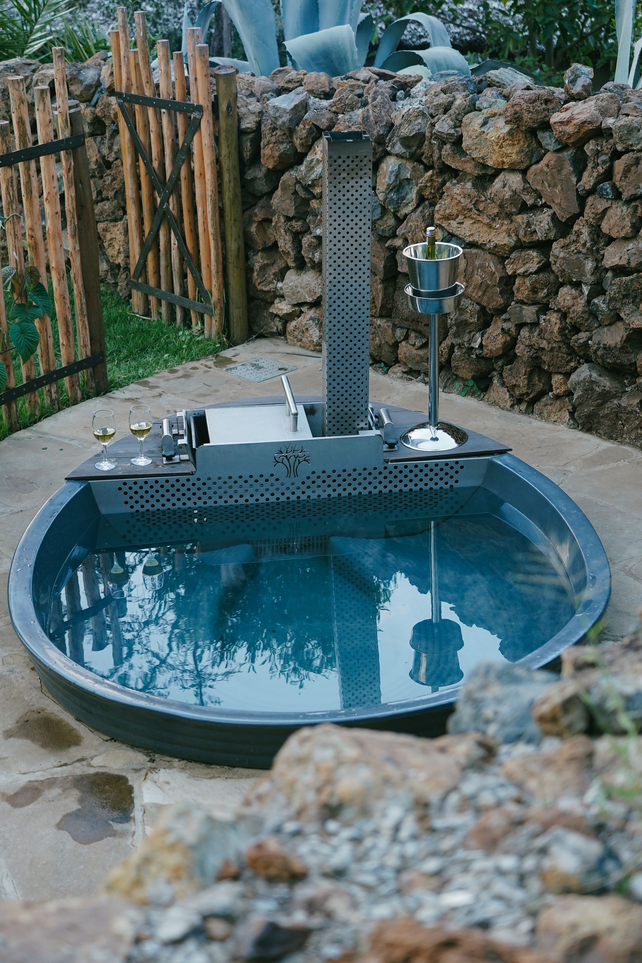The round wood-fired tub against a dry-stone wall and agave, two glasses of white wine and an ice bucket at its edge.