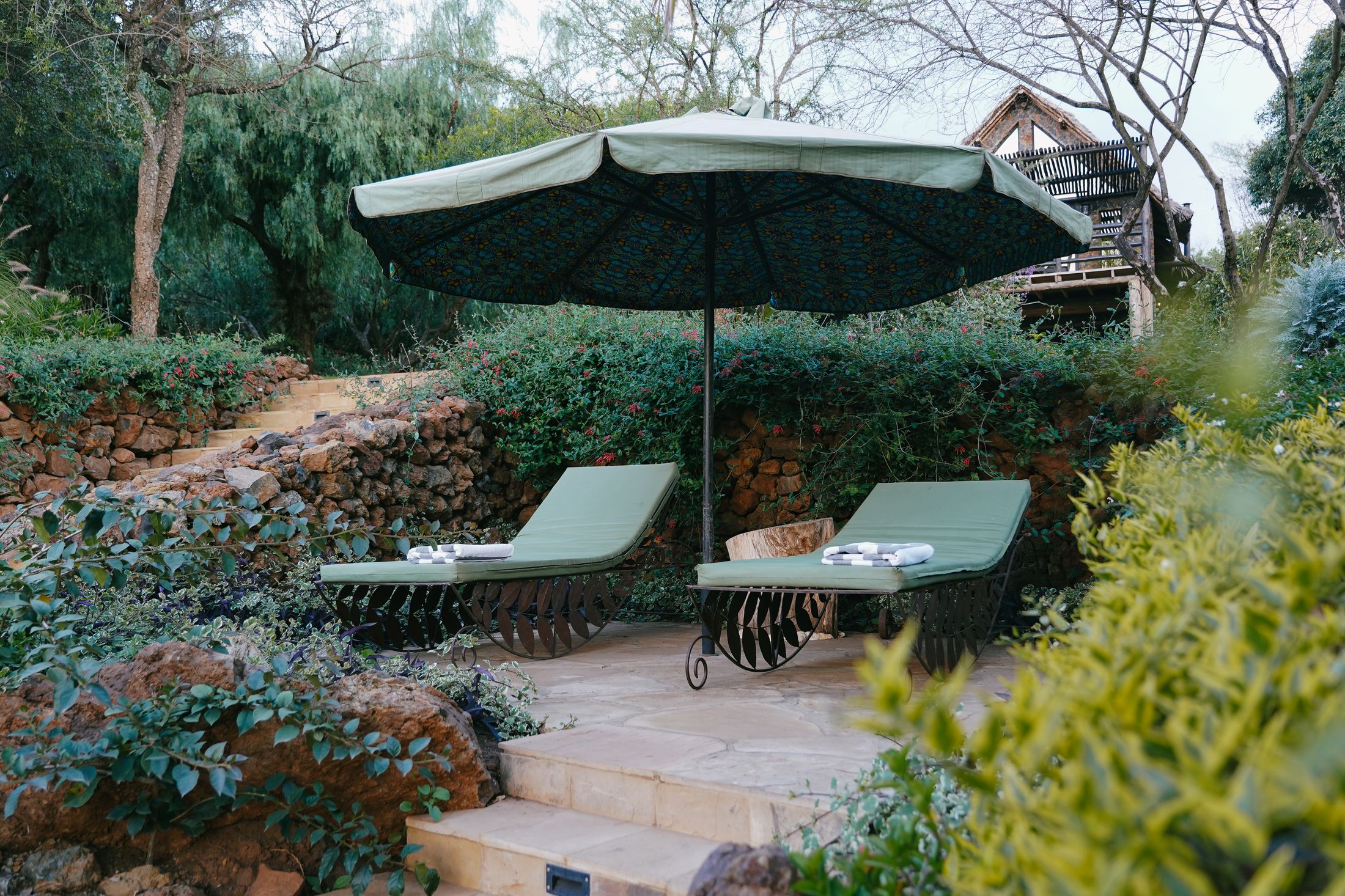 Two loungers under a kanga-lined umbrella on a flagstone landing, lava-rock terrace walls and stone steps rising behind through the planting.