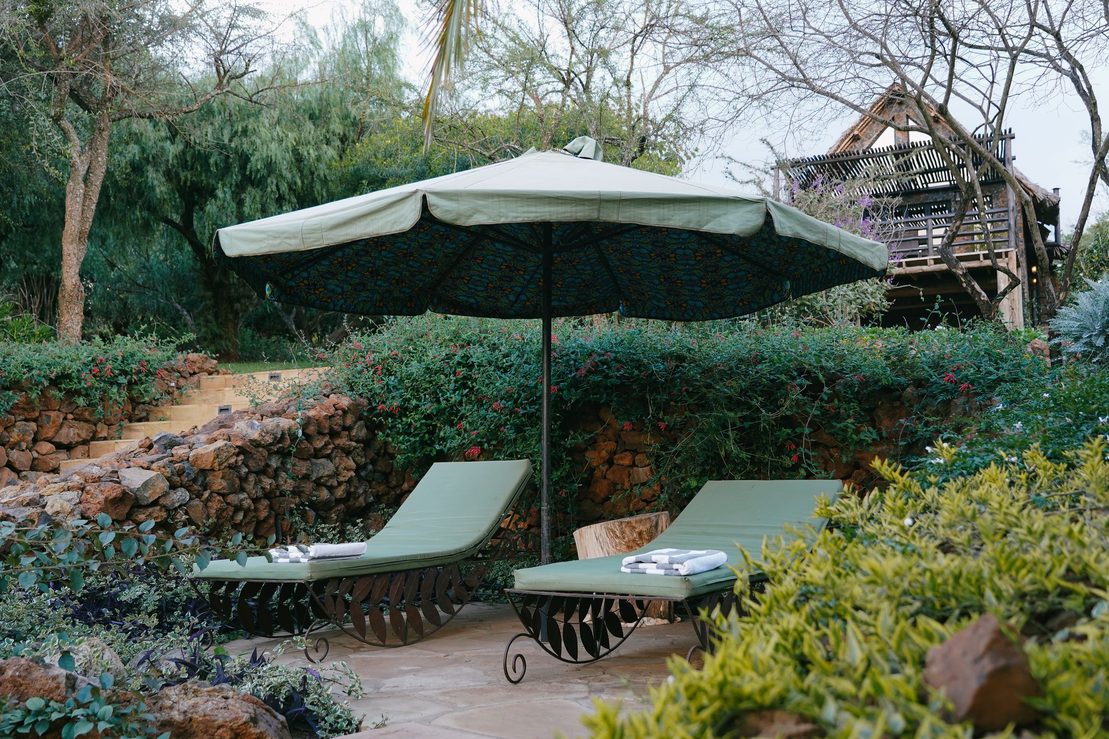 Two loungers under a garden umbrella beside a low stone wall, the thatched deck of O'Lorien behind the trees.