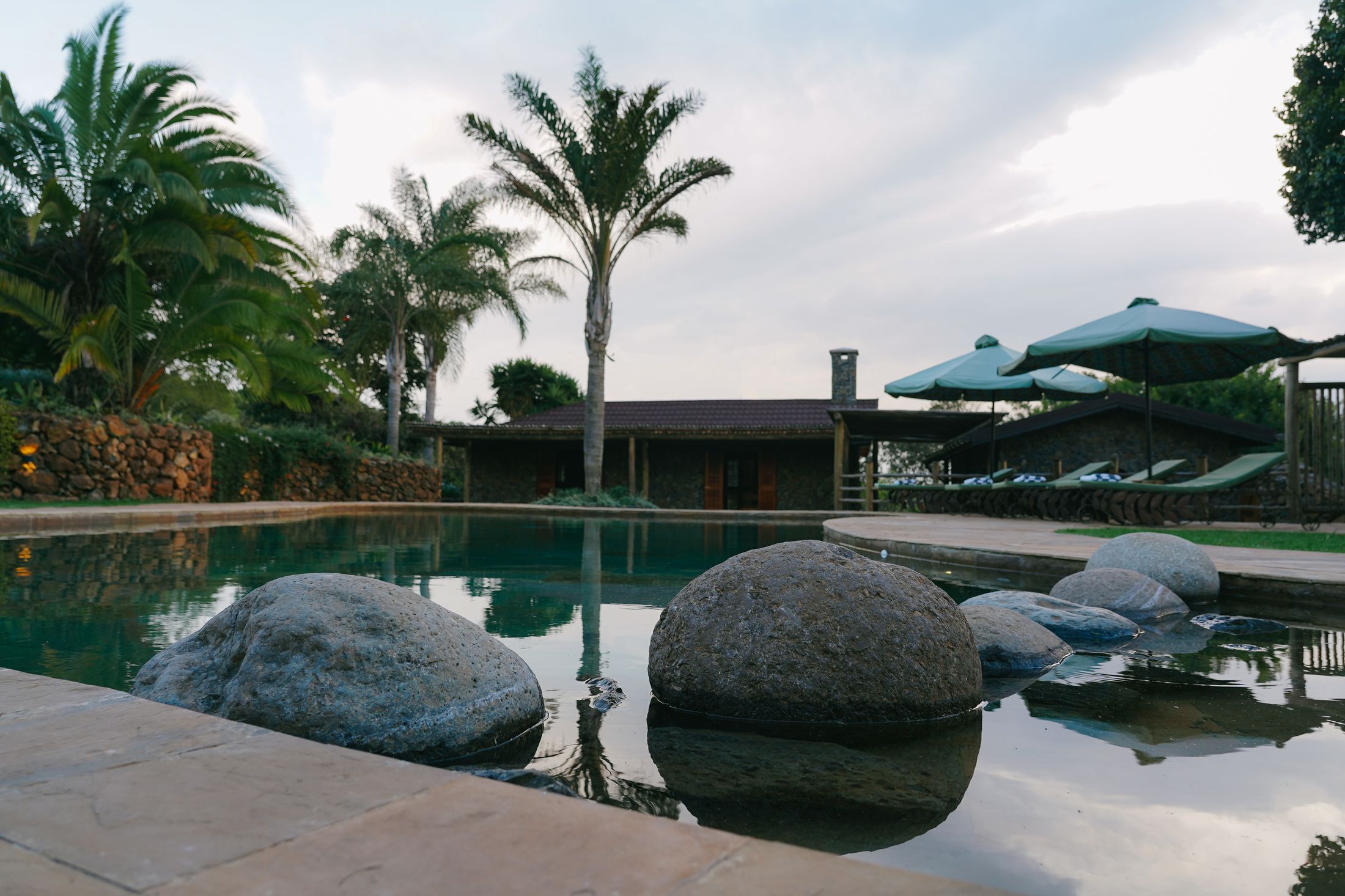 The free-form pool under an overcast sky, boulder stepping stones breaking the still water, palms and the stone pool house beyond.