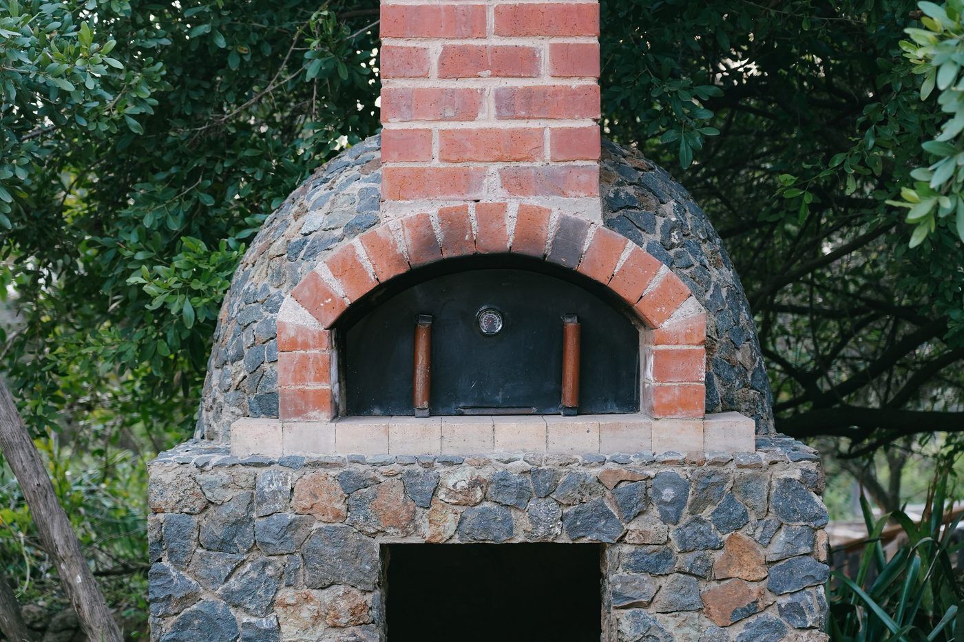 A stone and red-brick wood-fired pizza oven with an arched mouth, standing among garden foliage.