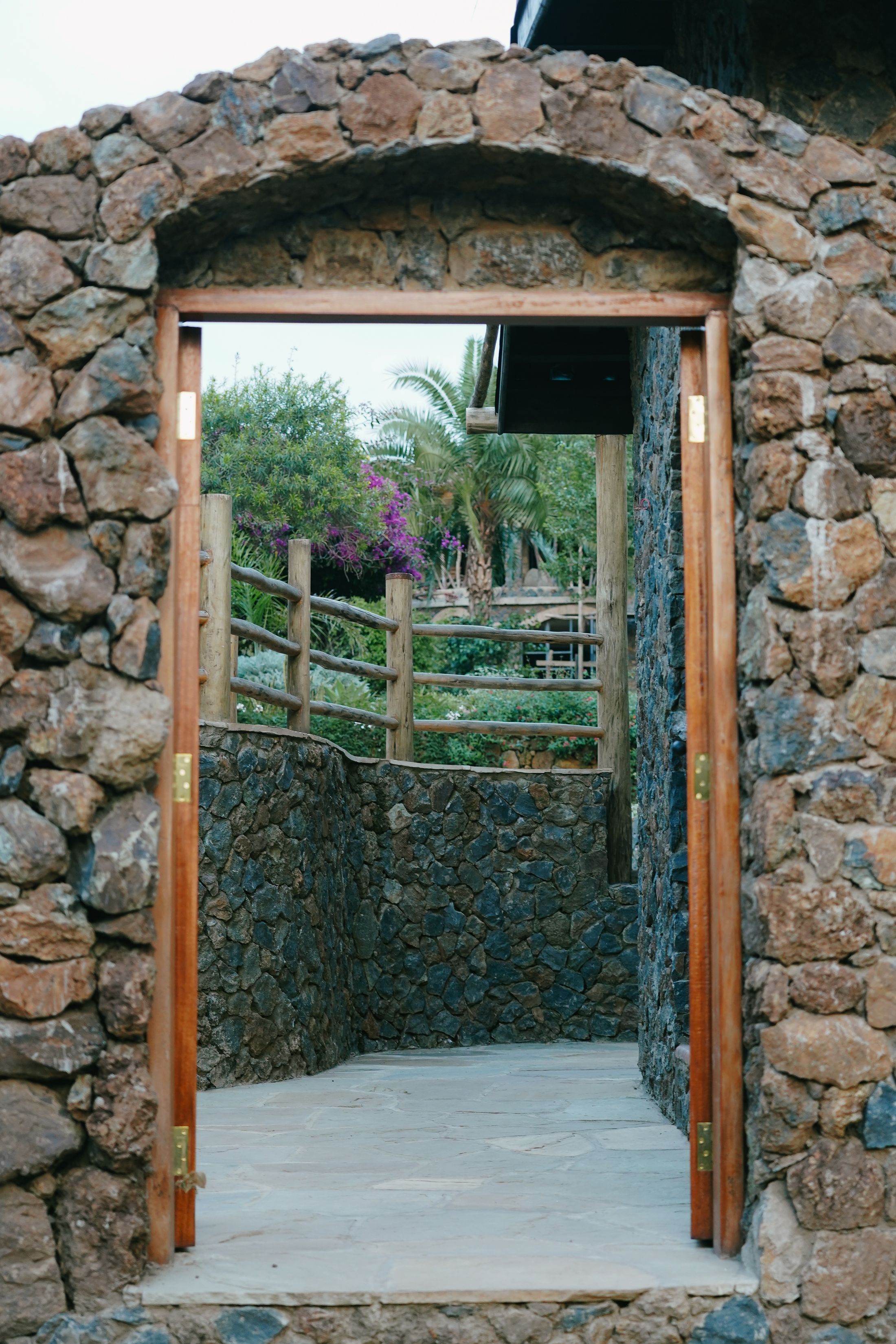 A stone arch doorway opening onto a walled garden path, a log-rail gate, bougainvillea and a palm beyond.