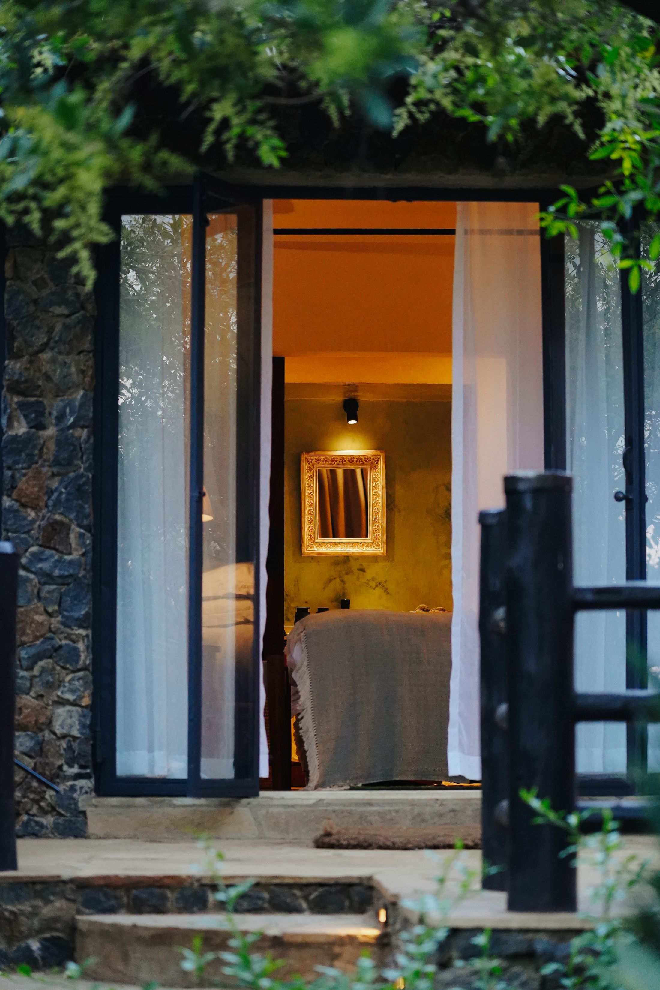 Dusk view through the open glazed doors of a stone cottage into a lamplit bedroom — green plaster walls, a gilt mirror, the bed turned down.